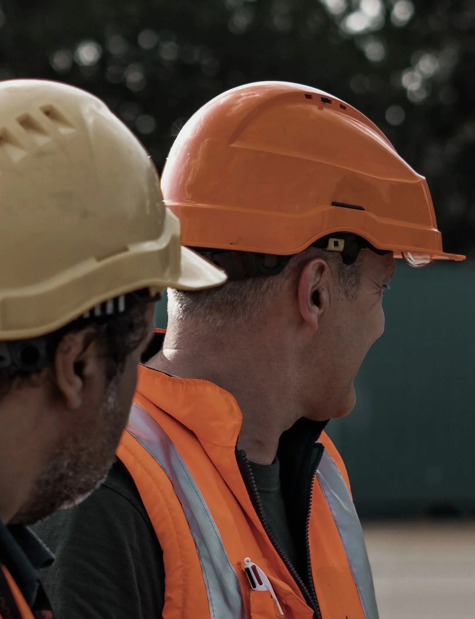 Close-up image of two construction workers wearing safety helmets and orange safety vests, with the focus on the helmets and the side of one worker's face.