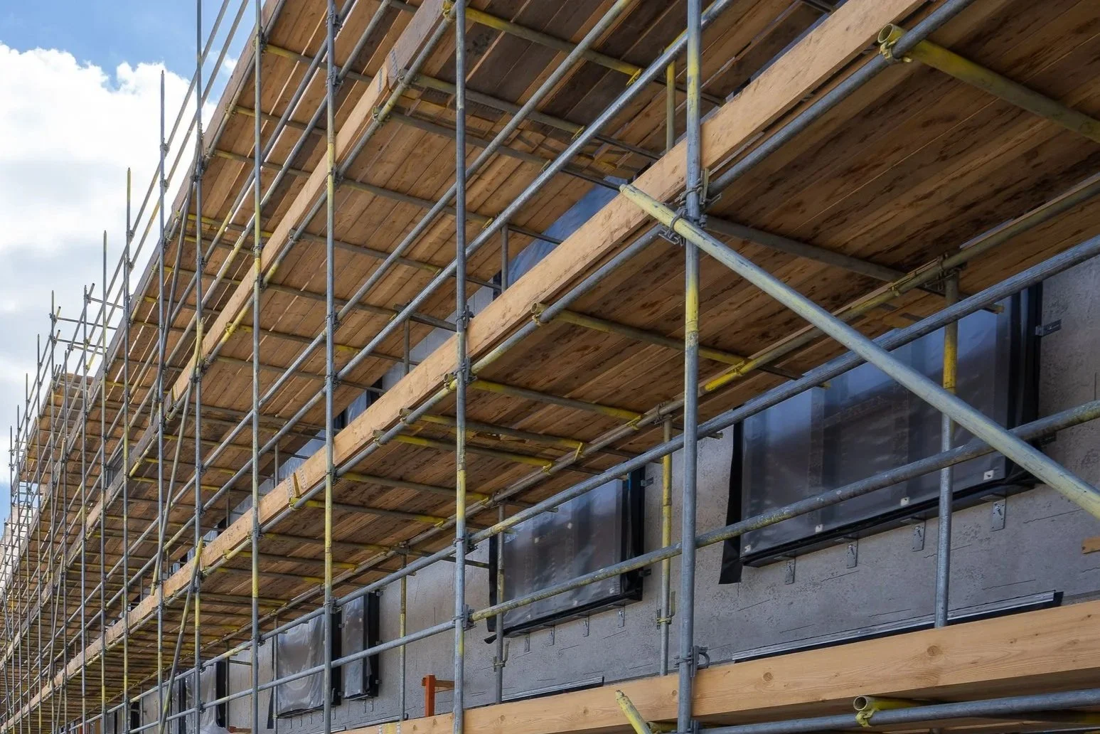 Construction scaffolding around a building with visible windows, partially covered with plastic sheeting.