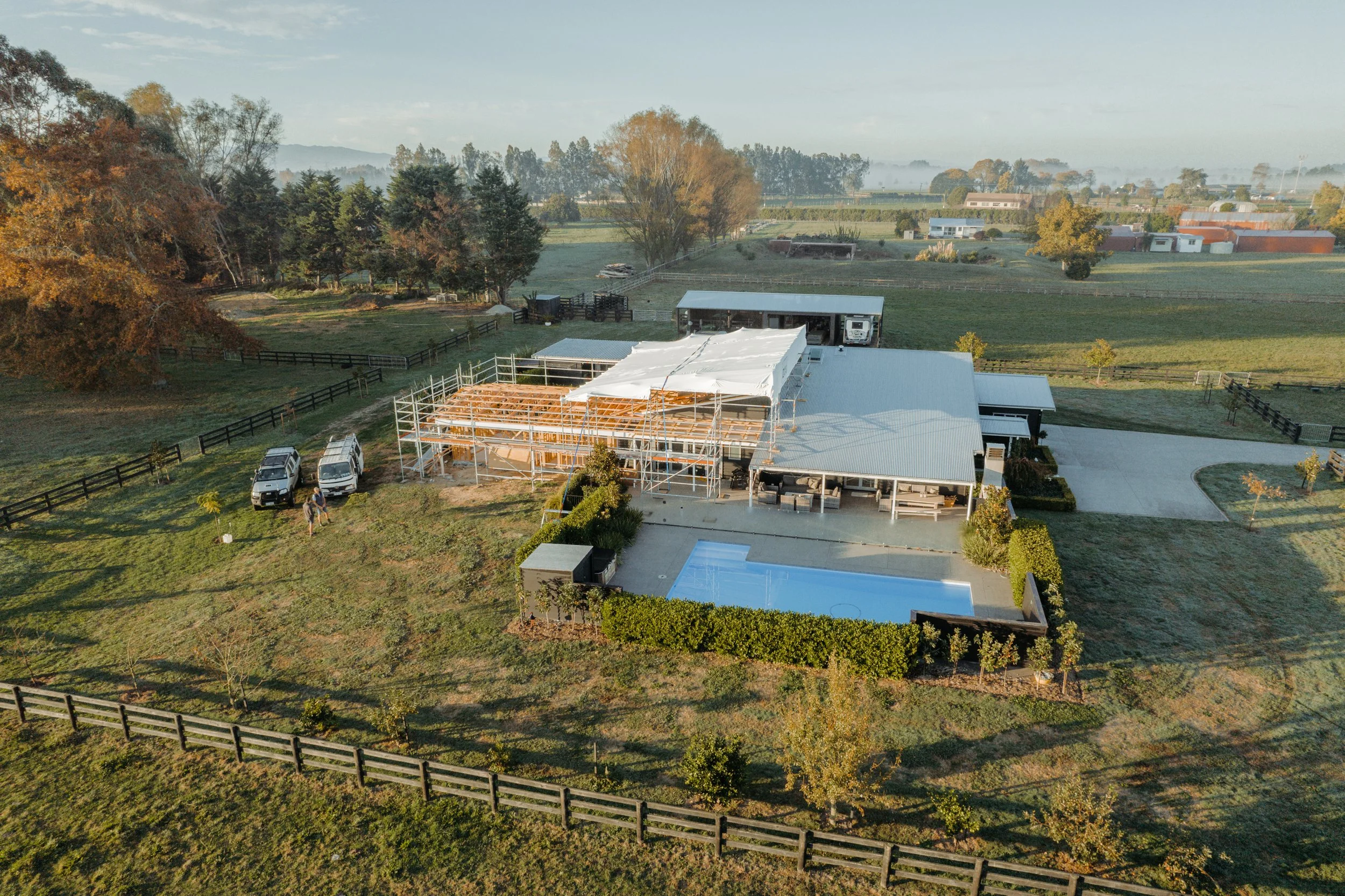 A Waikato New Zealand house under construction, scaffolding, a swimming pool, and multiple trees with autumn foliage. There are two vehicles parked next to the house, and farm buildings and fields extend into the distance.