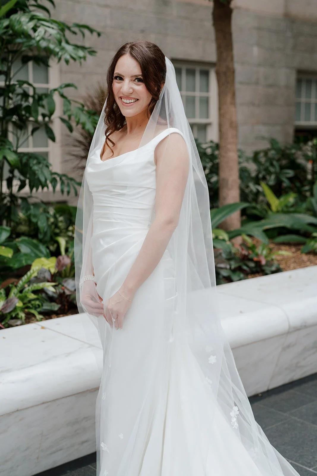 A smiling woman wearing a white bridal gown and veil standing outdoors near greenery and a stone building.