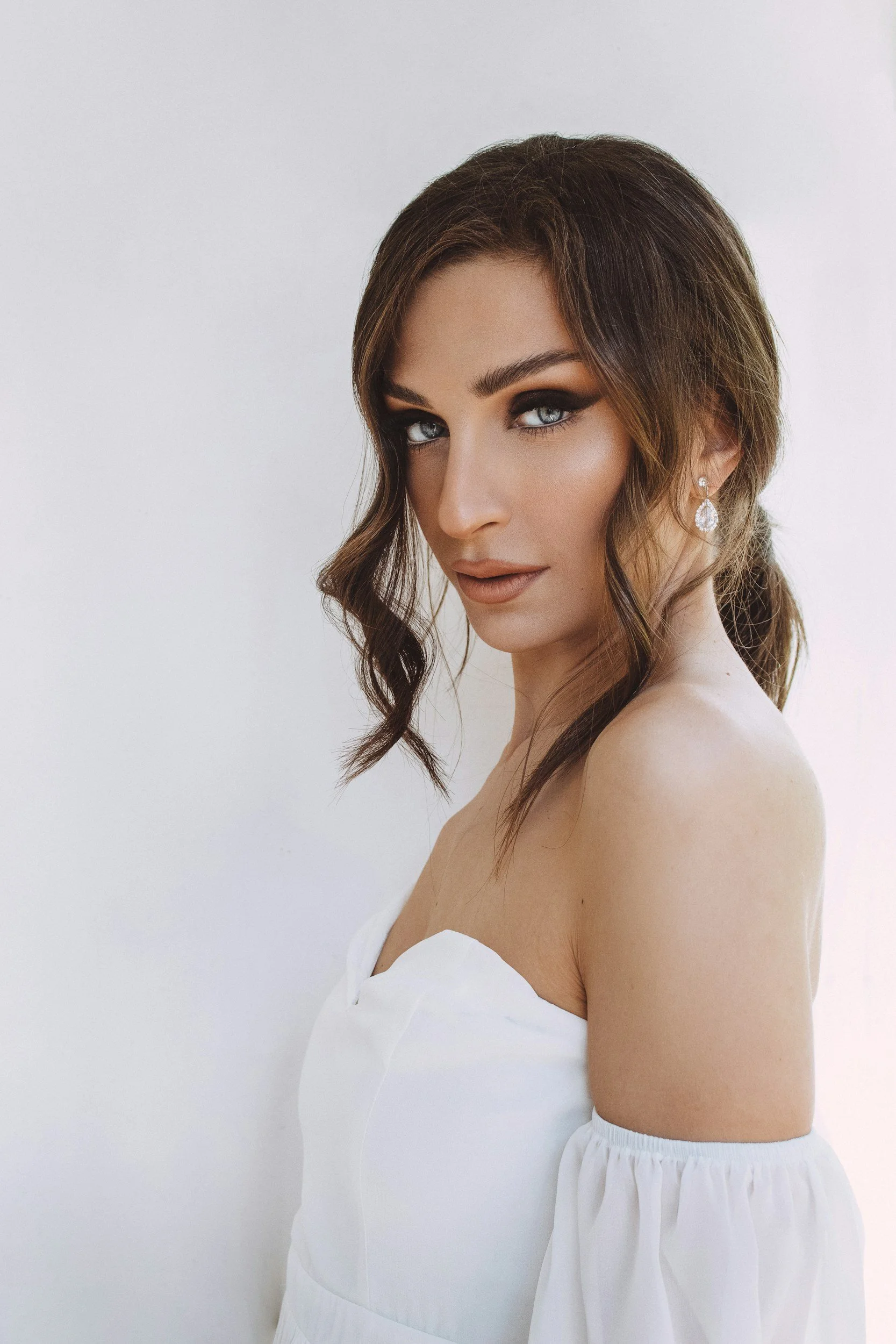 A woman with wavy brown hair, blue eyes, and makeup, wearing earrings and a white off-shoulder dress, posing against a plain background.