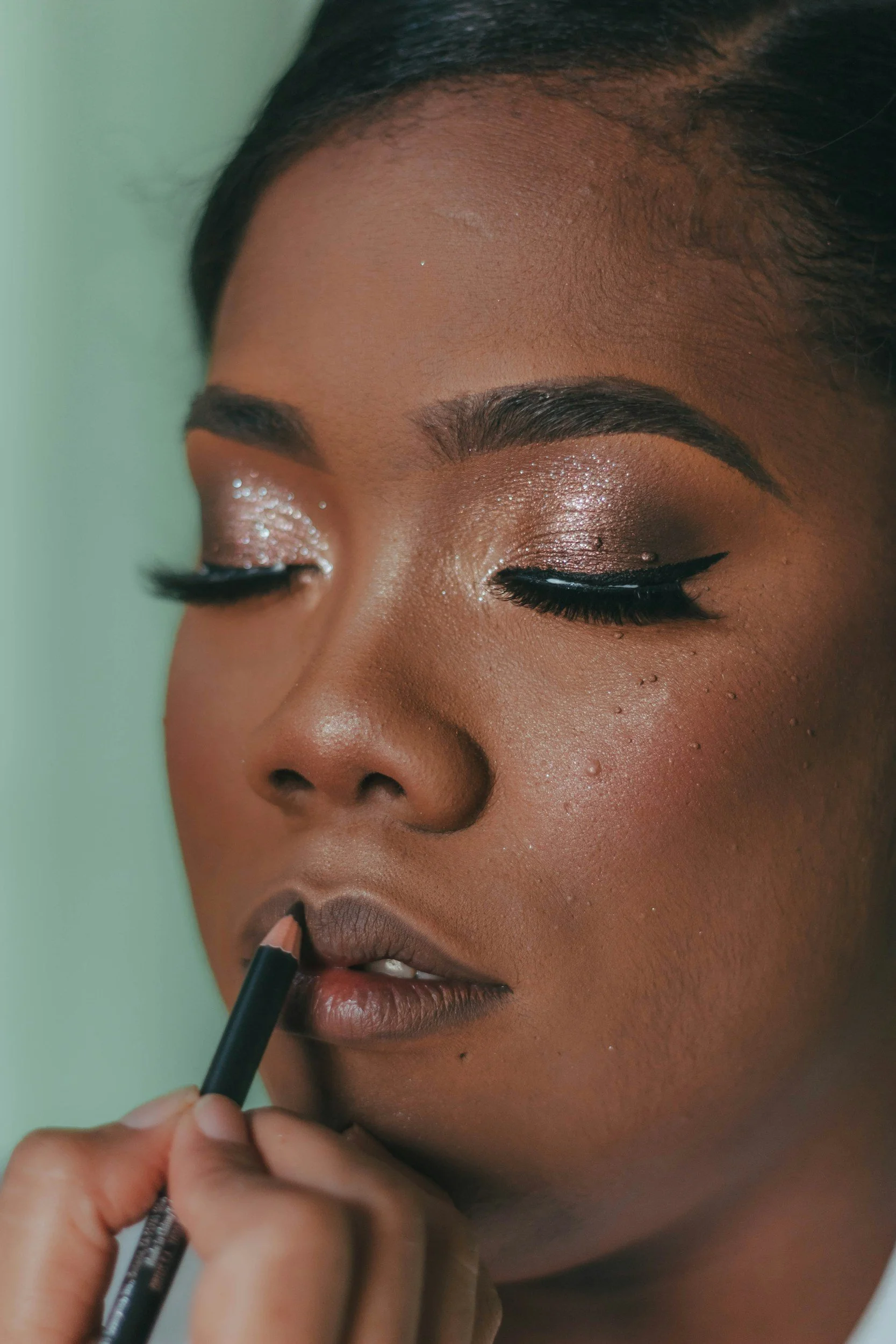 Close-up of a woman with dark skin applying lip liner, her eye makeup includes shimmery eyeshadow and winged eyeliner, and her eyebrows are well-defined.