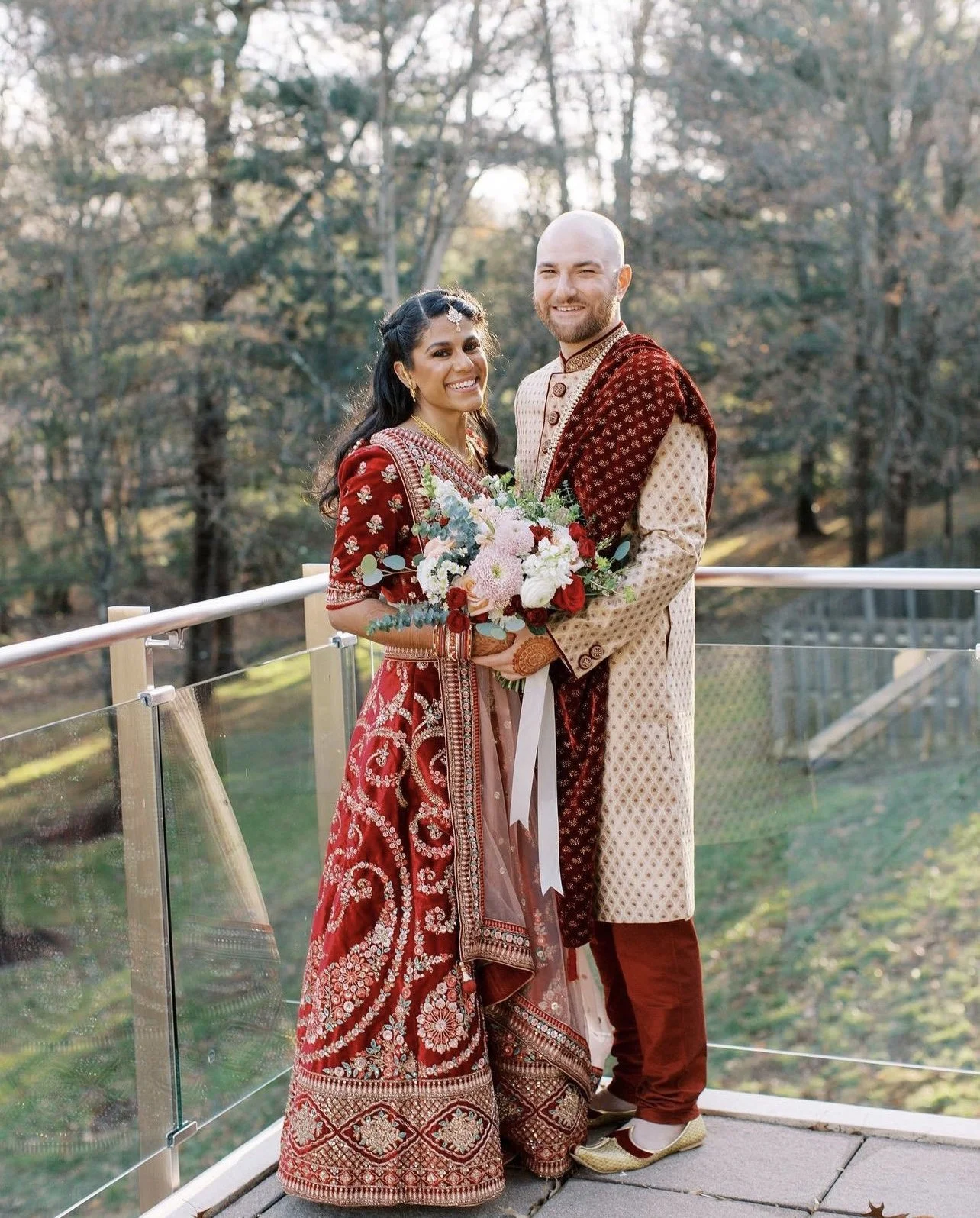 A newlywed couple in traditional Indian attire standing outdoors on a balcony, smiling and holding a bouquet of flowers with trees and natural scenery in the background.