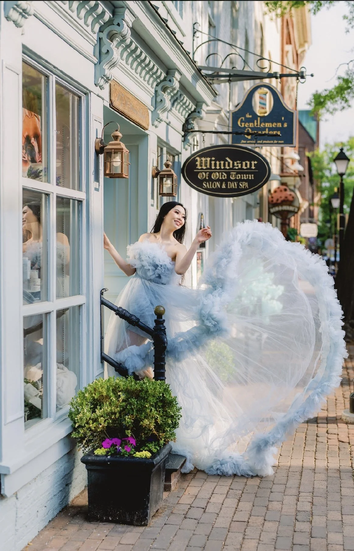 A woman in a white, fluffy dress is posing on a town sidewalk outside a storefront, holding a large parasol. She is smiling and appears to be enjoying a sunny day.