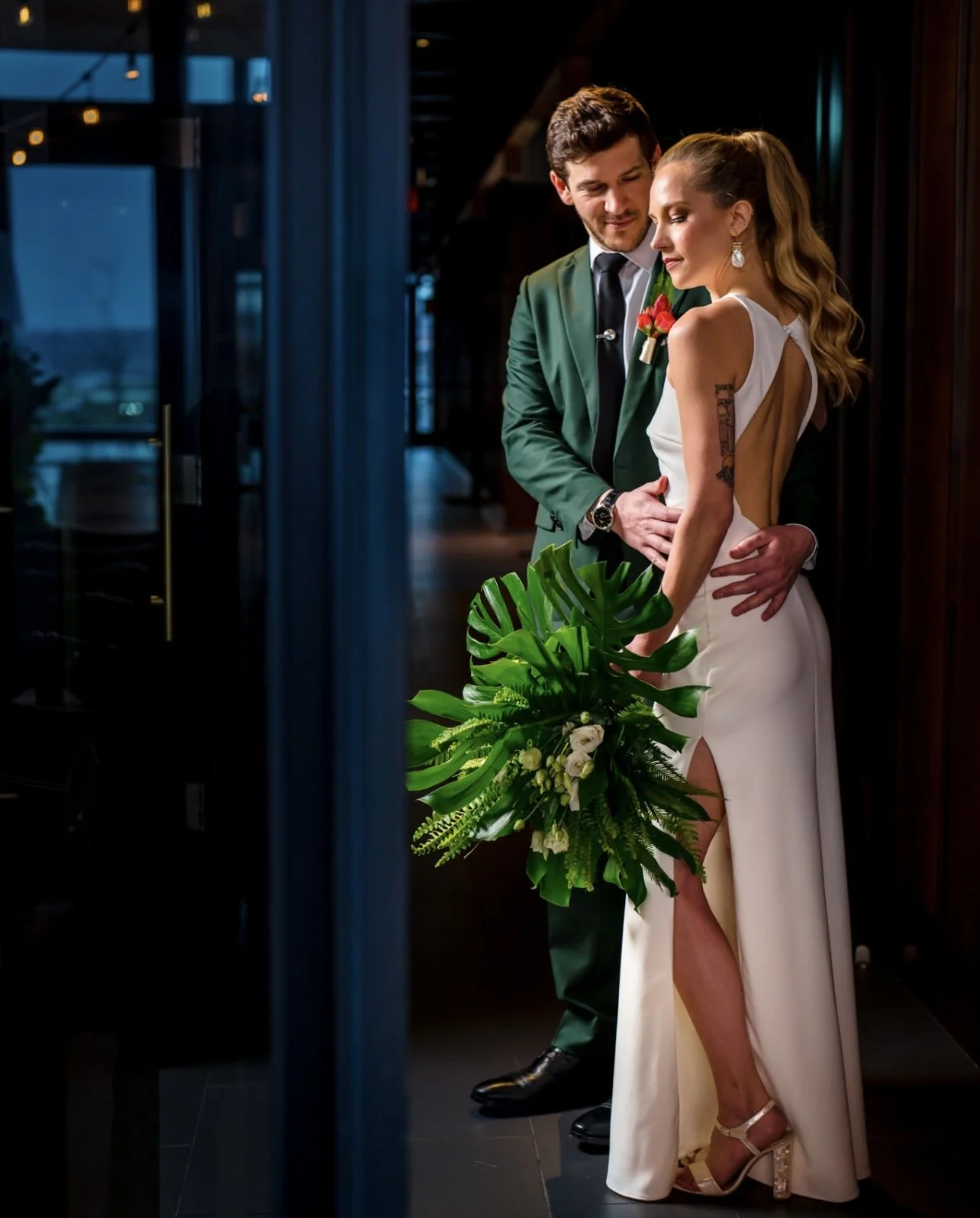 A bride and groom sharing a romantic moment, with the groom gently holding the bride's waist as she holds a large bouquet of green leaves and white flowers. The bride is wearing a white gown with a high slit, and the groom is dressed in a dark green suit with a black tie. They are standing indoors near a dark wooden wall, with soft lighting creating an intimate atmosphere.