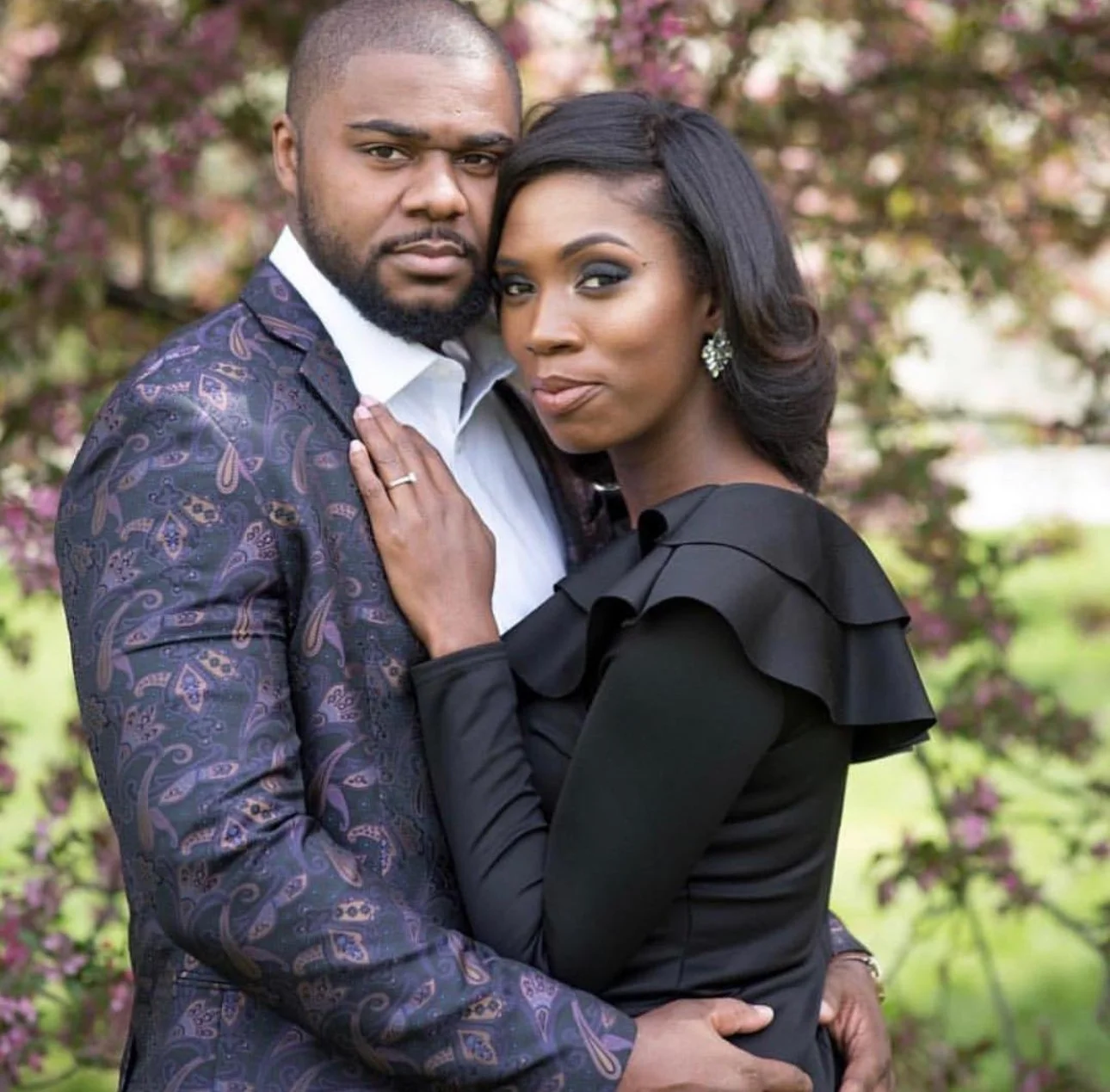 A couple posing outdoors during springtime, standing close and looking at the camera. The man has a beard and is wearing a purple patterned suit jacket over a white shirt. The woman has long dark hair styled in waves, wearing earrings and a black dress with ruffle details on the shoulders. The background is blurred with pink blossoms and greenery.