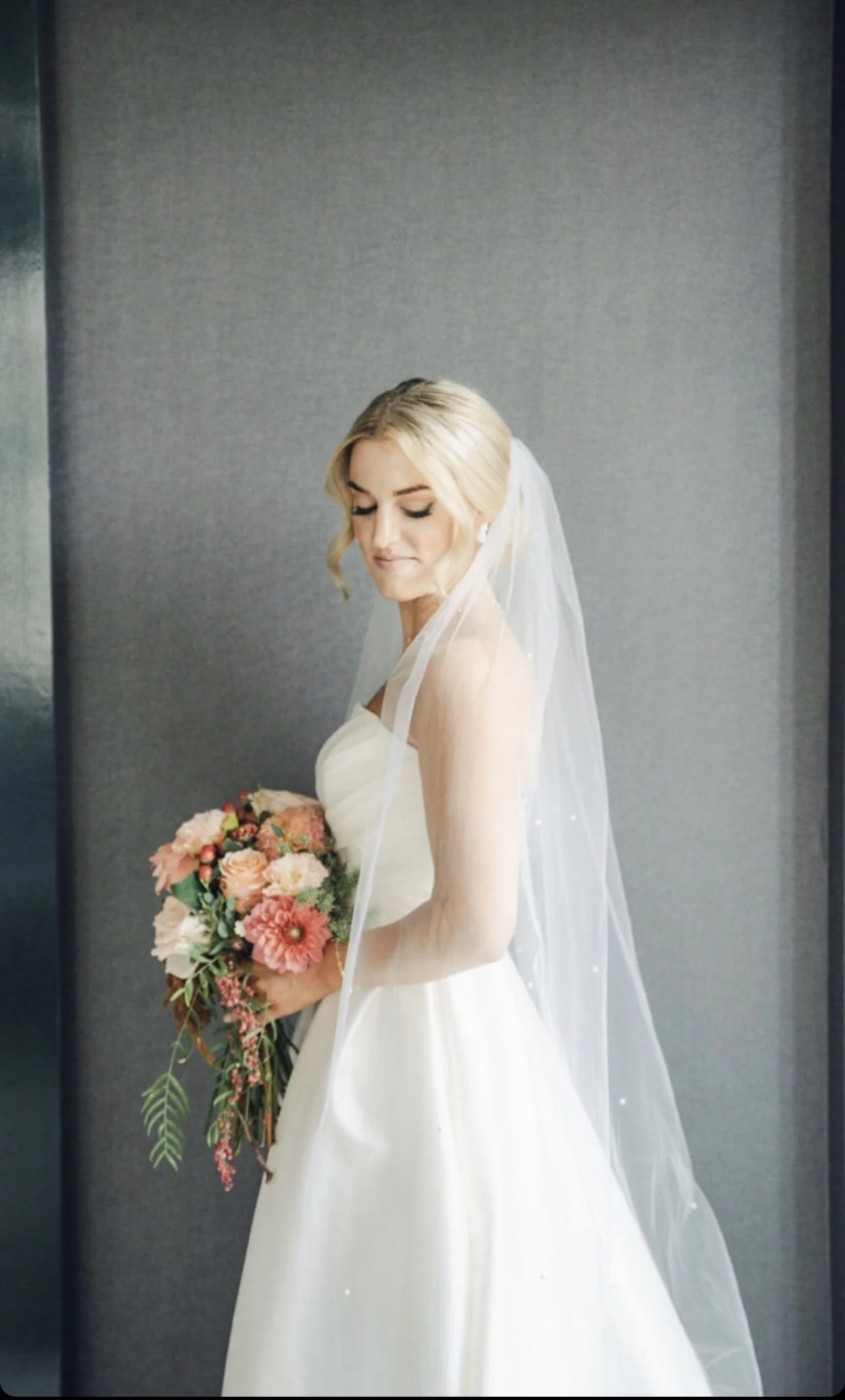 A bride in a white wedding dress holding a bouquet of pink and peach flowers, with a sheer veil over her blonde curls, standing against a plain grey wall.