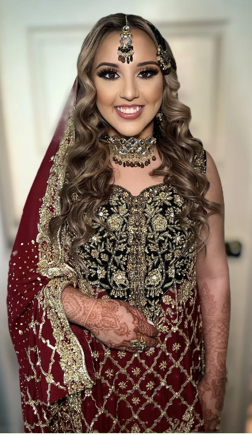 A woman dressed in traditional South Asian attire with intricate gold and black embroidery, wearing jewelry including a necklace, earrings, and forehead ornament, and decorated hands with henna.