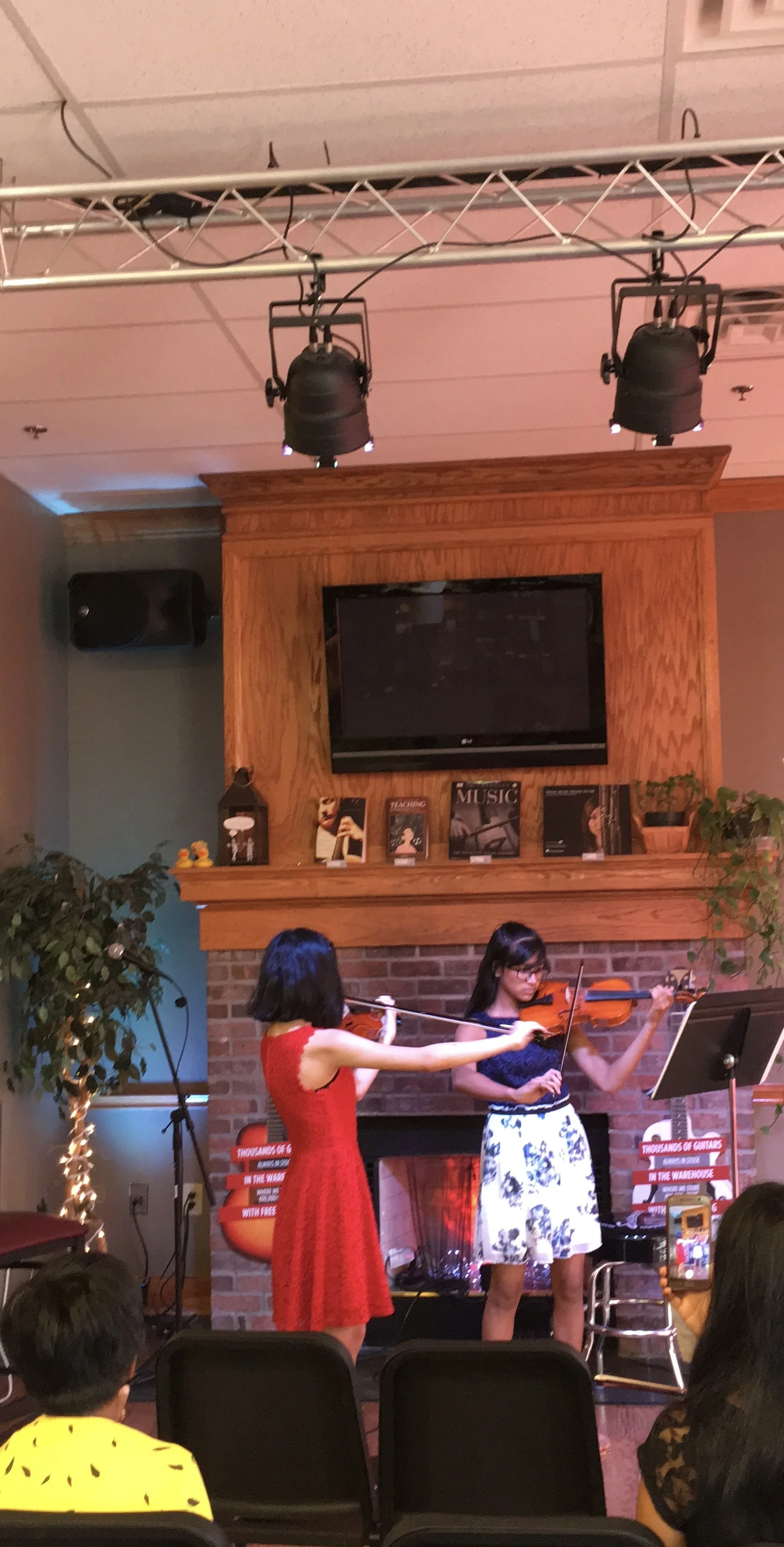 Two women playing violins in a room with a brick fireplace and music-themed decor; audience members are watching and recording.