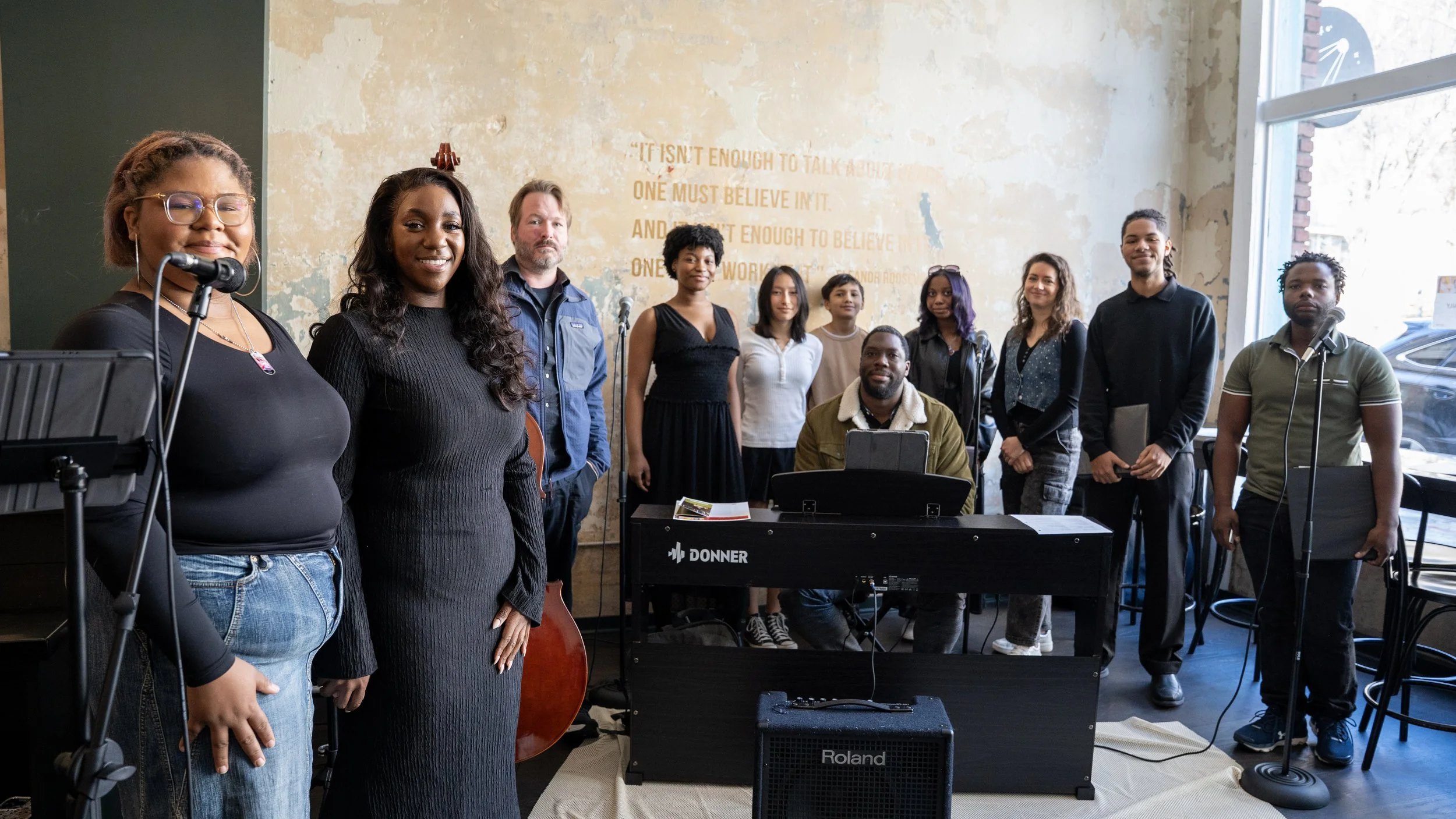 Group of diverse people gathered in a room, some holding musical instruments and others standing behind a keyboard, with a large window to the right and a partially worn wall with quote art in the background.