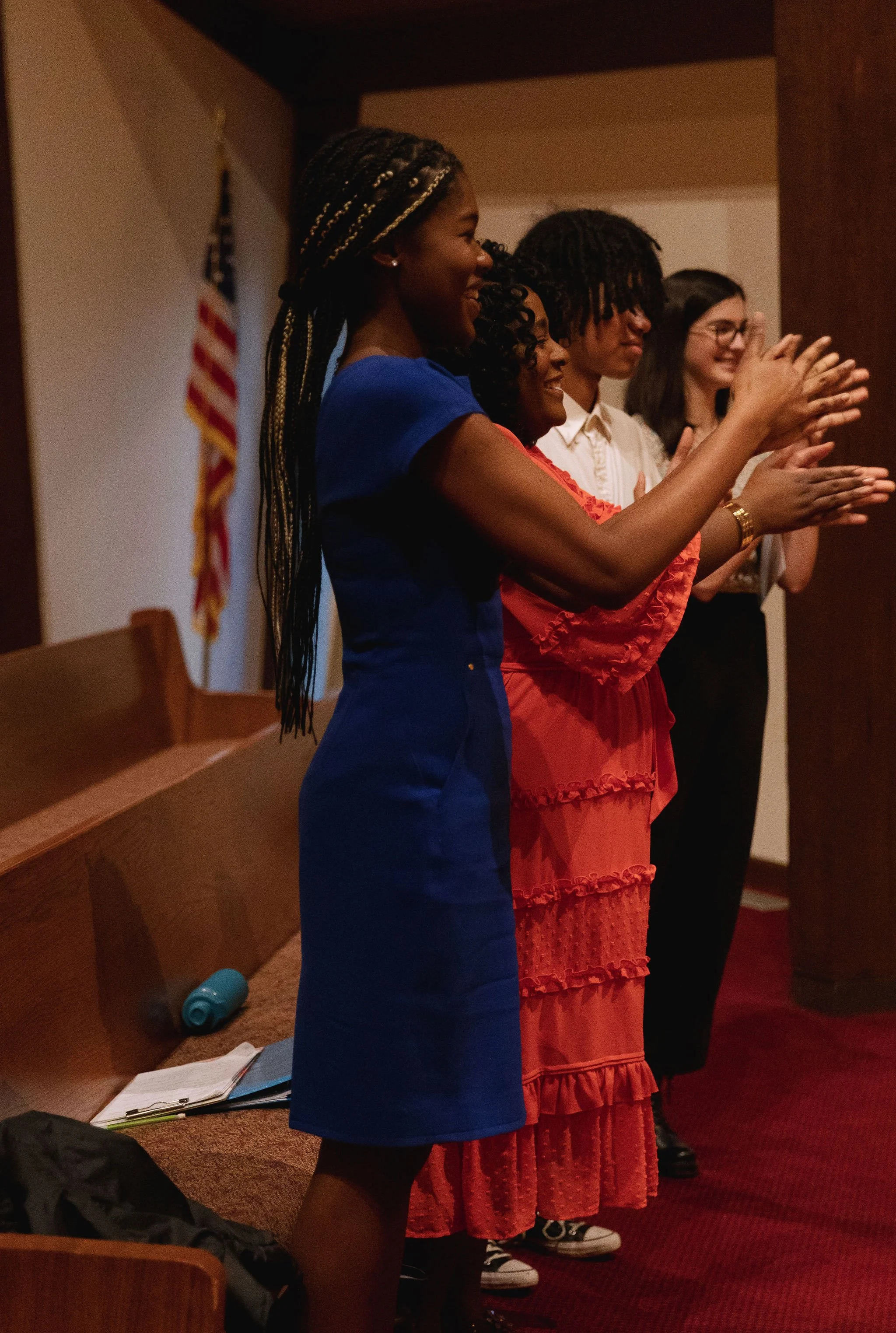 Group of people clapping in a room with an American flag in the background, standing near pews.