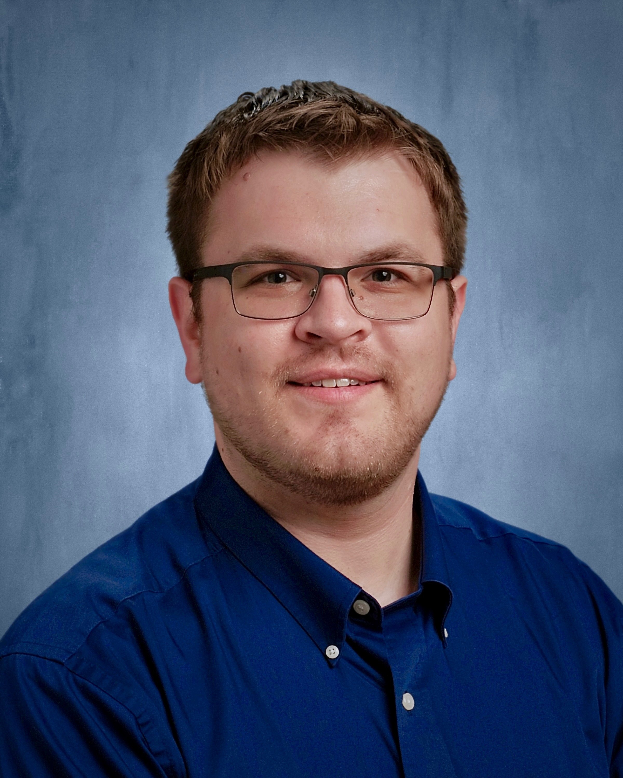 A young man with short brown hair, glasses, and a light beard wearing a blue collared shirt, smiling in front of a blue gradient background.