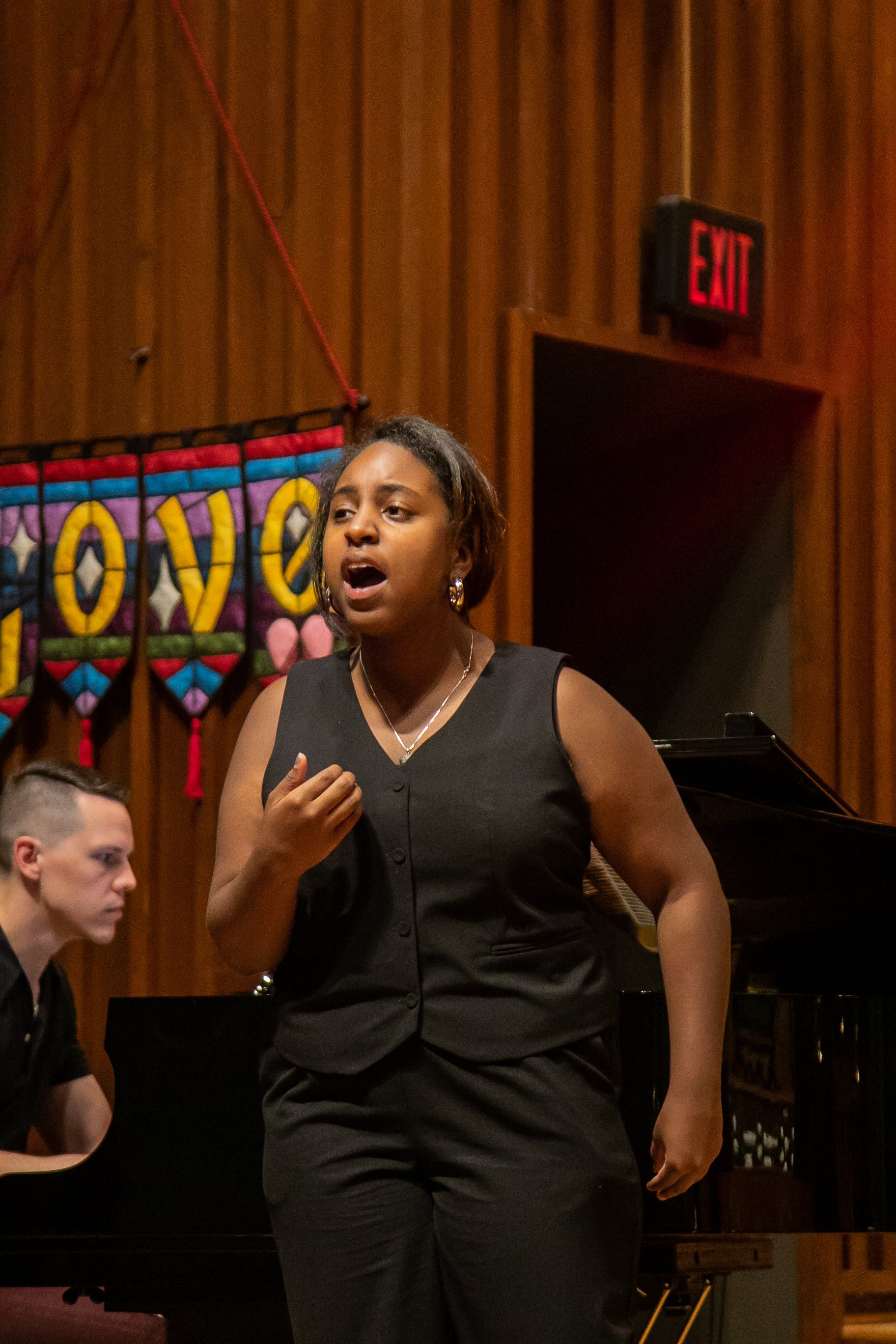 A woman singing passionately in a church with a pianist accompanying her. A colorful banner reading 'LOVE' hangs in the background, and an 'EXIT' sign is visible.