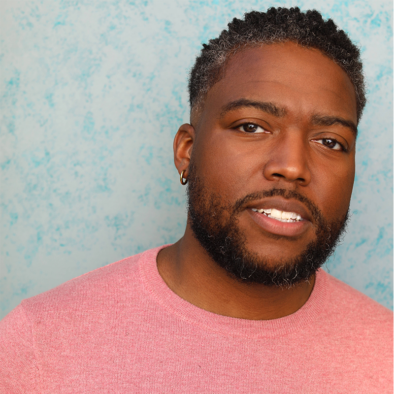 A man with short curly hair and a beard, wearing a pink sweater and small hoop earring, standing in front of a light blue textured background.