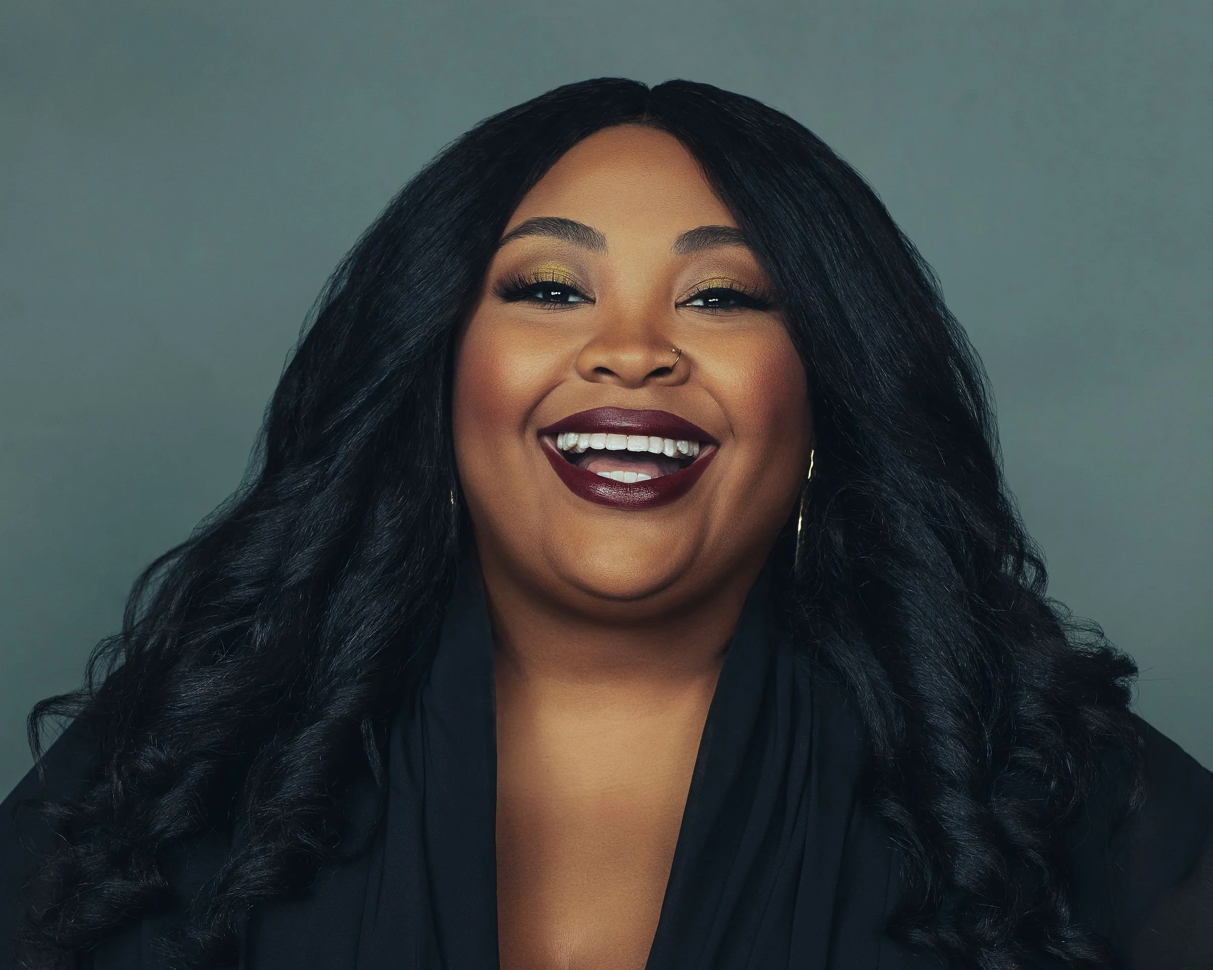 A woman with long, wavy black hair and dark lipstick smiling against a neutral background.