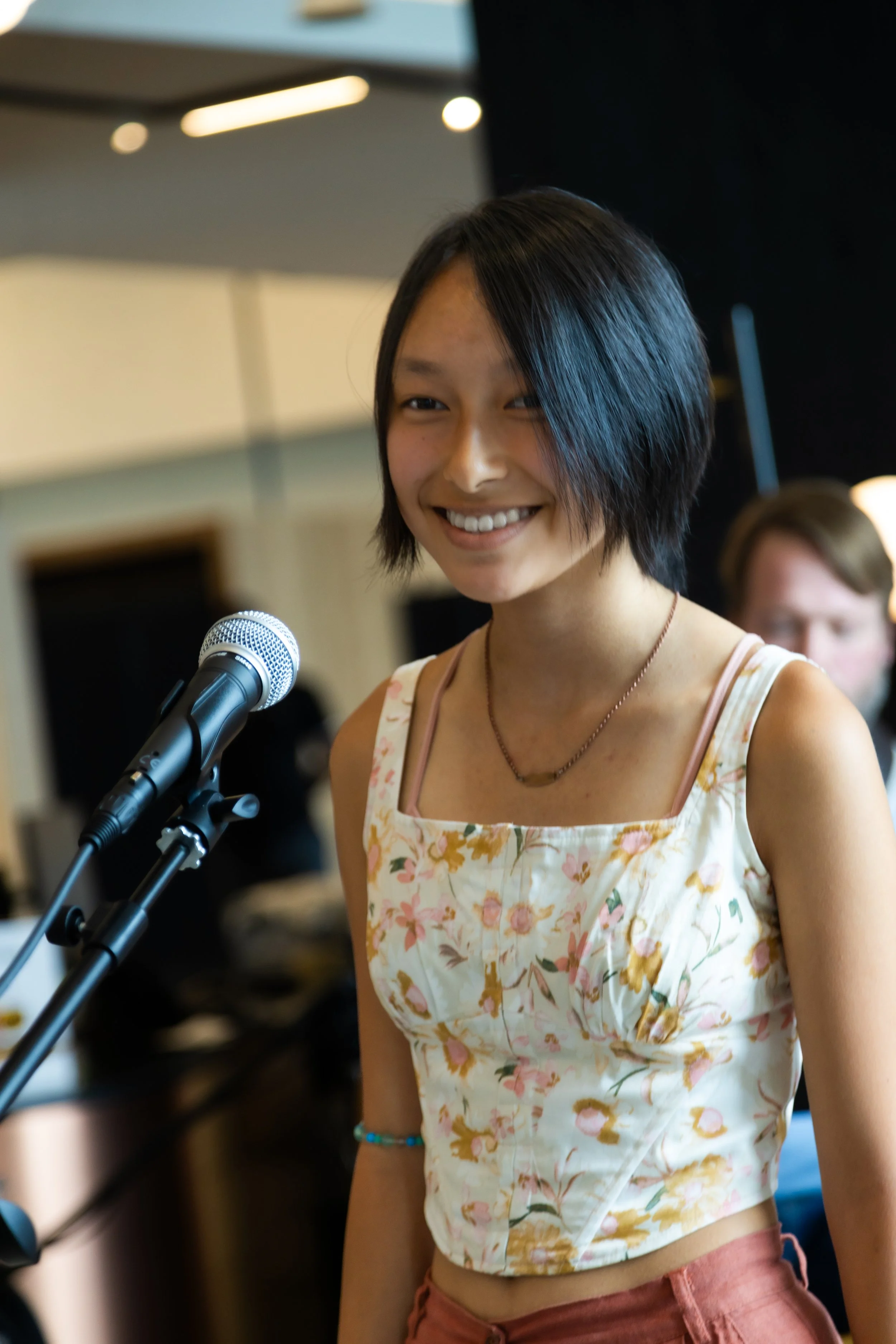 Person smiling in front of a microphone, wearing a floral tank top and red pants, indoors. Burke Music Academy concert.