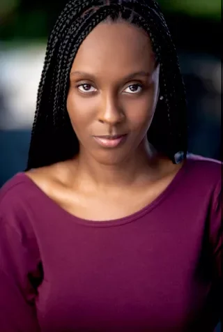 A young woman with braided hair wearing a purple top, looking directly at the camera with a neutral expression.