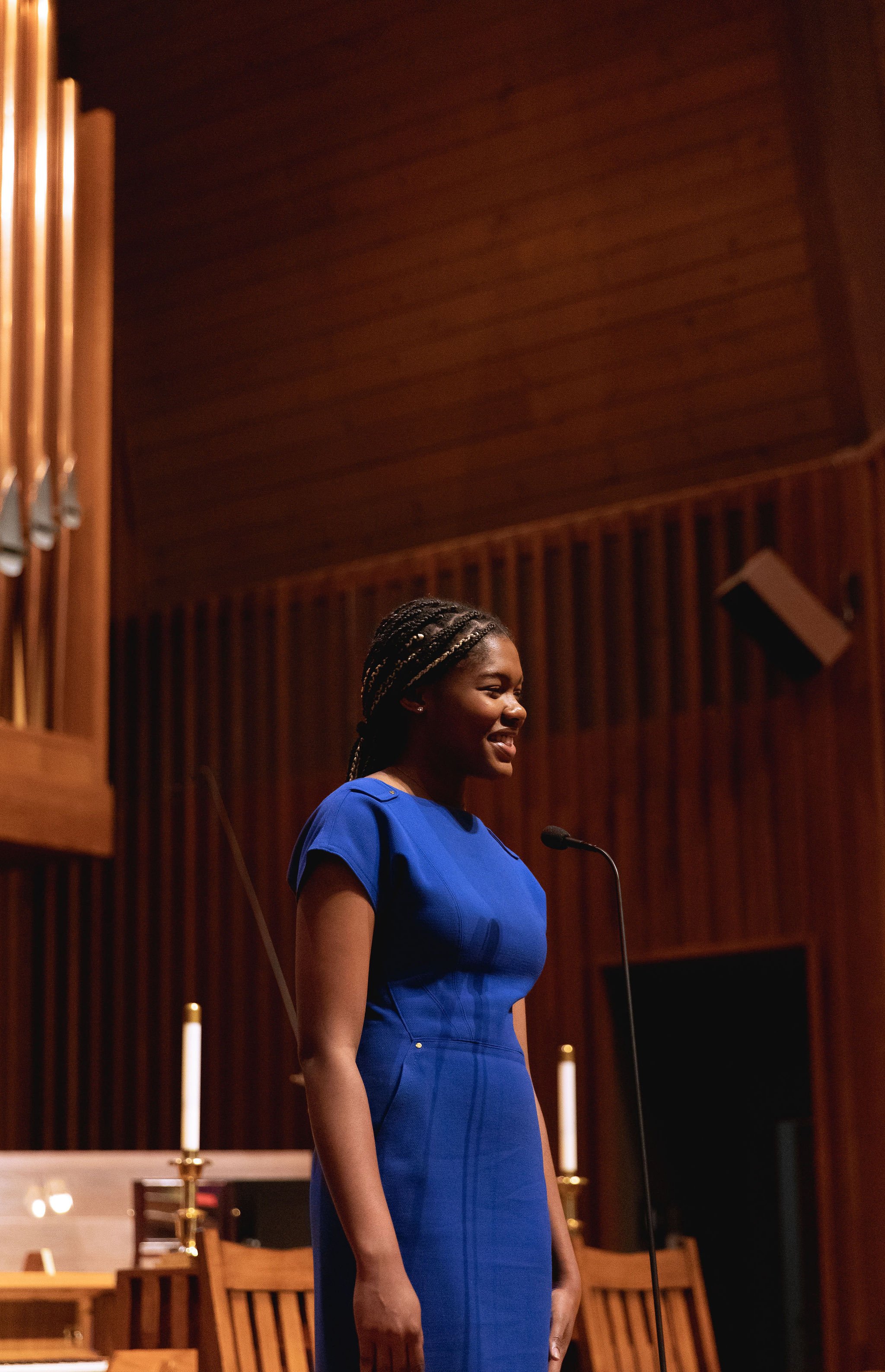 Woman in blue dress speaking at a microphone in a church with candles and organ pipes visible.