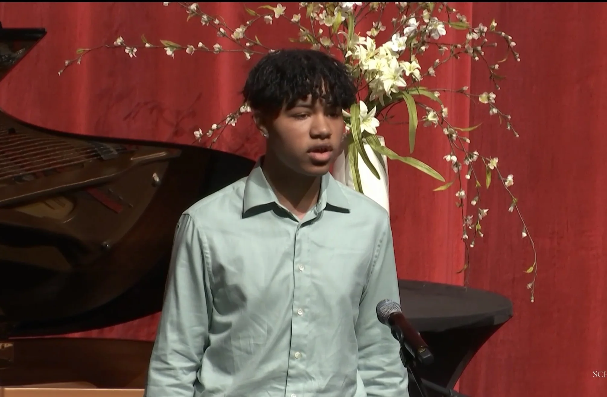 Young man in a light green shirt singing on stage with a microphone, red curtain background, grand piano, and floral arrangement.