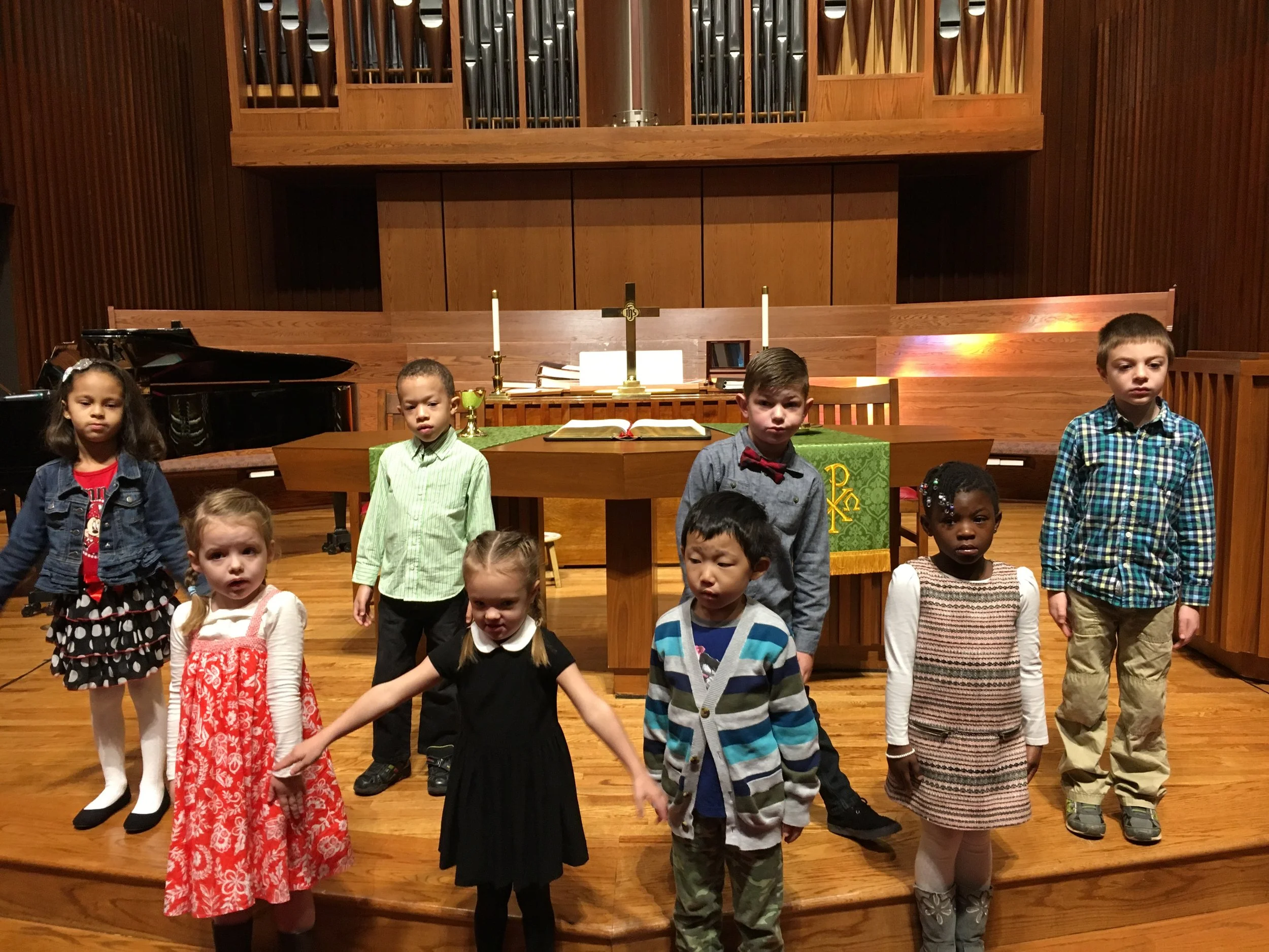 Children standing on a church altar, with a backdrop of a wooden pulpit, pipe organ, and piano. They are wearing a variety of colorful clothing, including dresses, sweaters, and a bow tie.