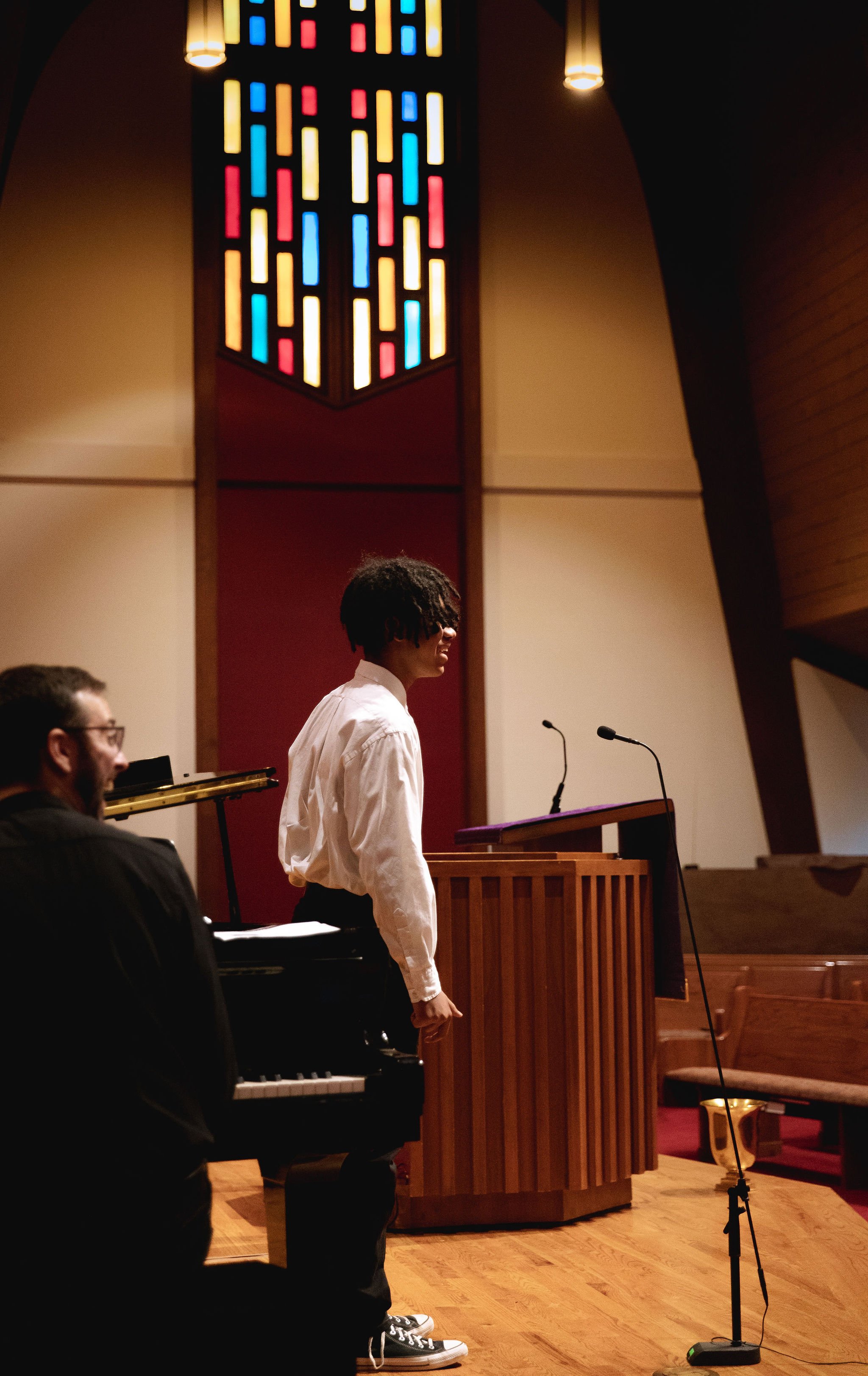 Two people in a church setting with a piano, wooden pulpit, and stained glass window.