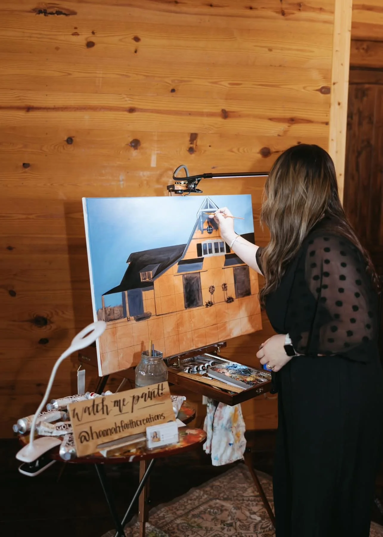 A woman painting a house with a blue sky background on a canvas. She is standing in a wooden room and appears to be in the process of creating a realistic architectural painting.