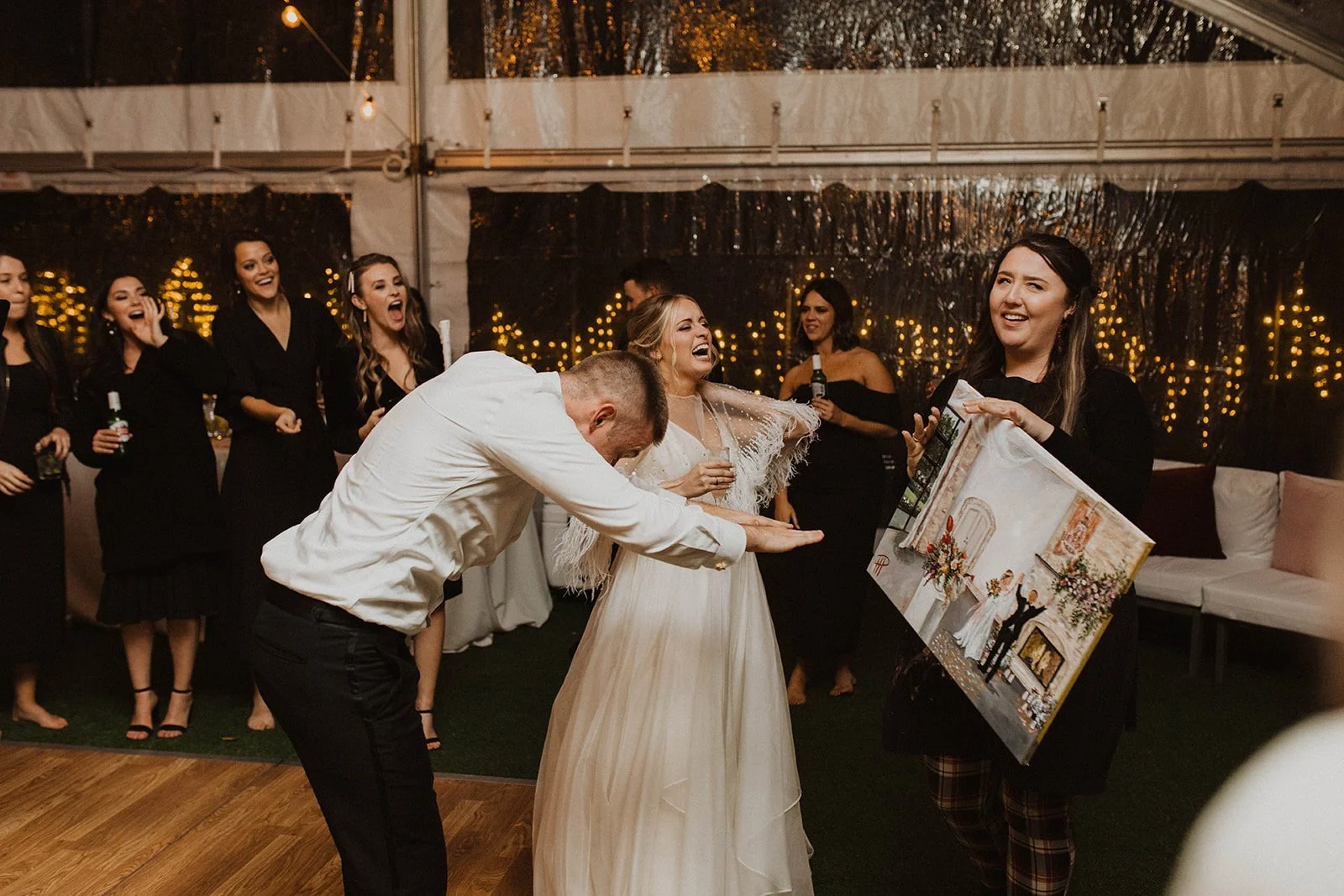 A wedding celebration with a group of women, including the bride in a white gown, laughing and smiling. A woman is holding a painting, and others are holding drinks. The scene is inside a tent with string lights