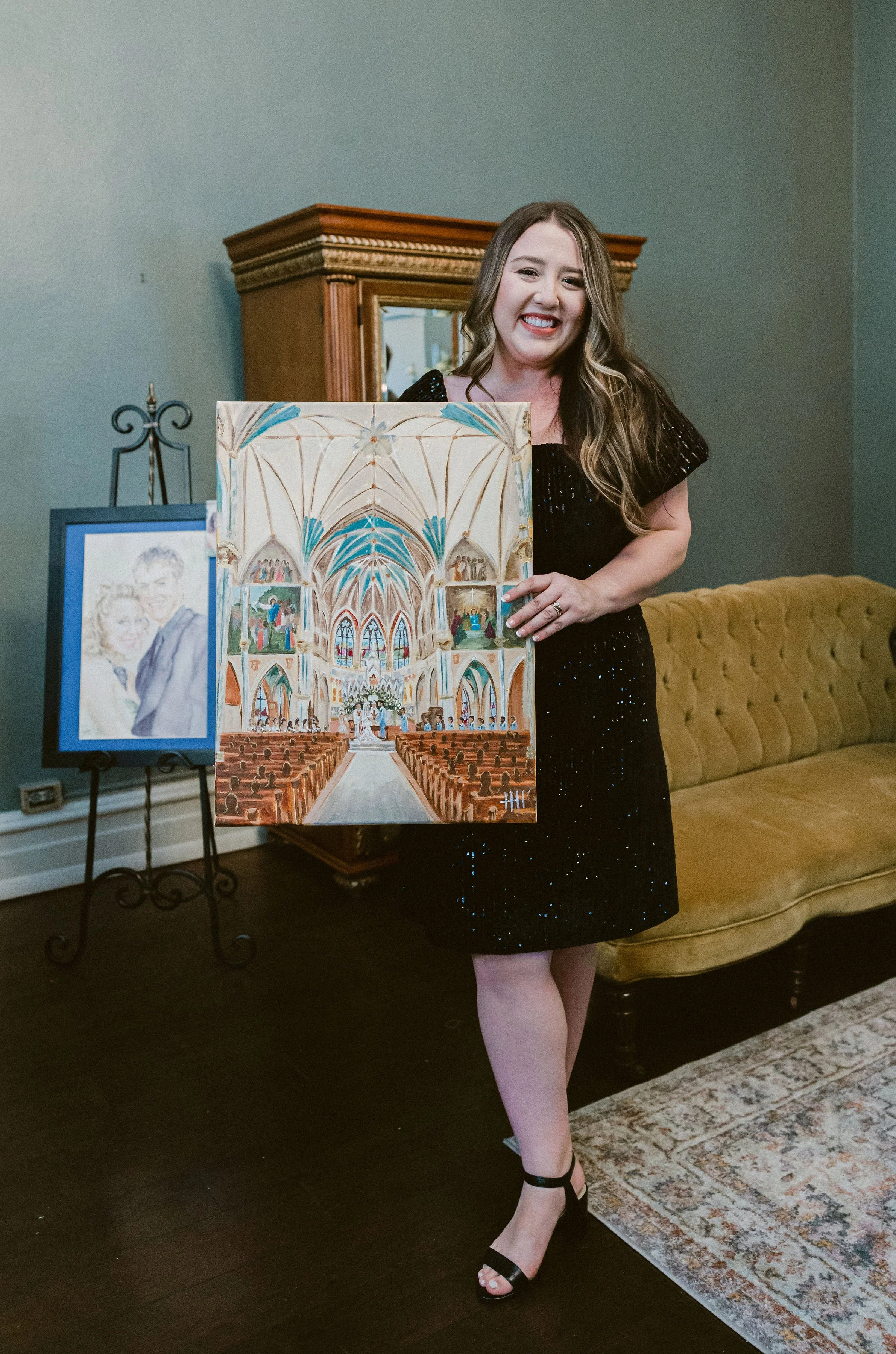 A woman in a black dress holding a painting of a church's interior during a social event, with an easel displaying a portrait of a couple and a velvet sofa in the background.