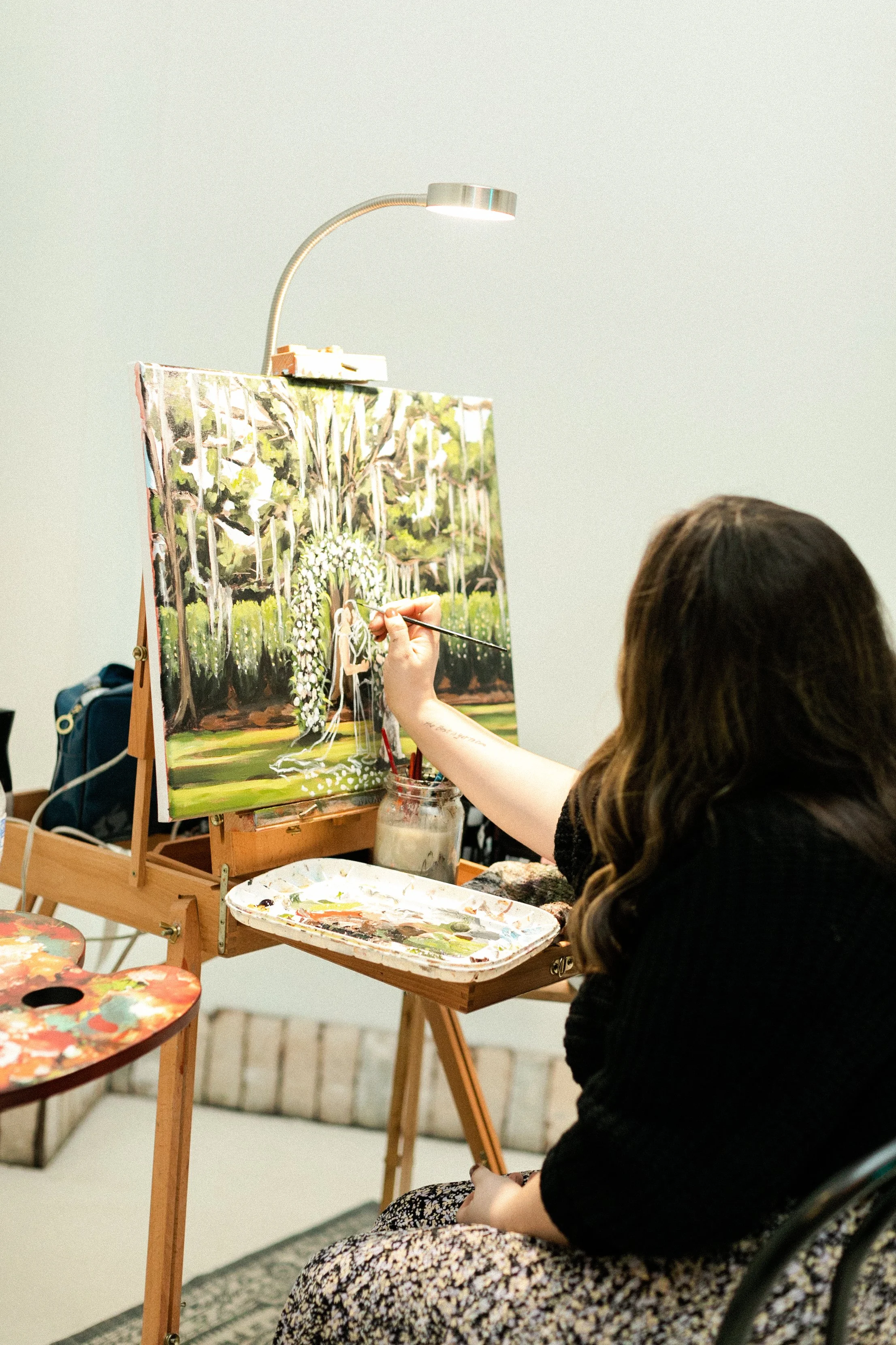 A woman painting a landscape scene on a canvas, sitting at an easel with painting supplies and a light fixed above.