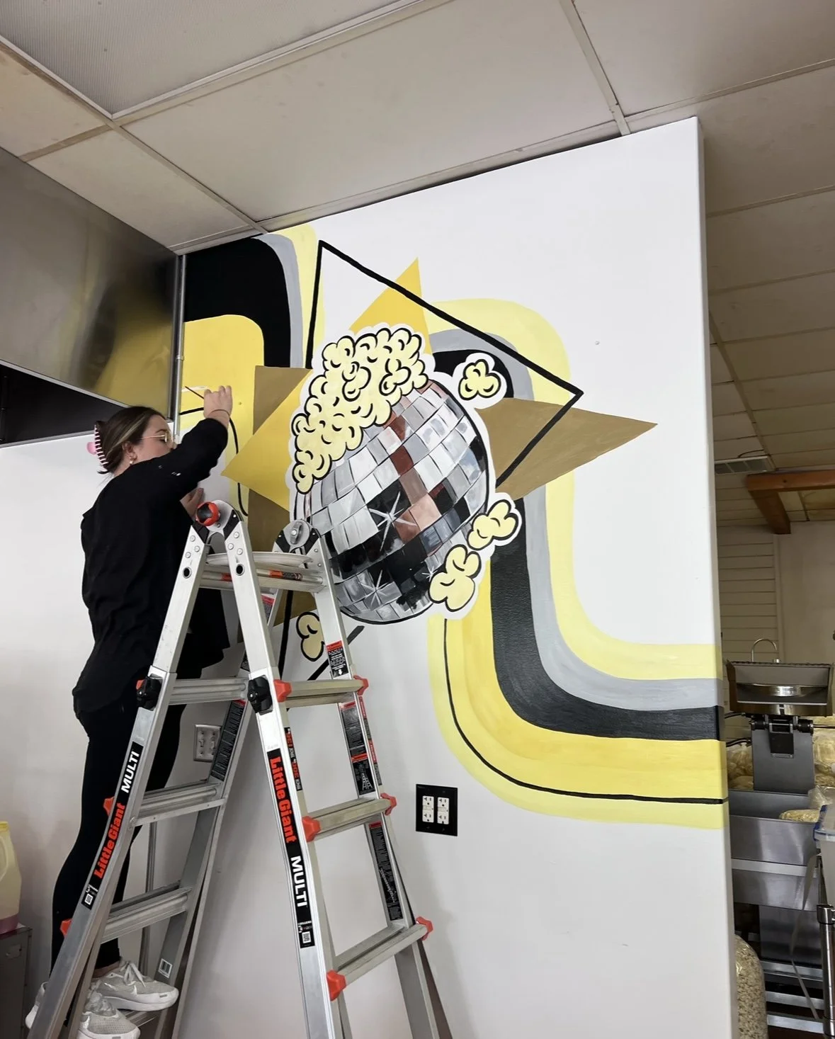 A person standing on a ladder painting a mural of a disco ball with popcorn and abstract shapes on a wall.