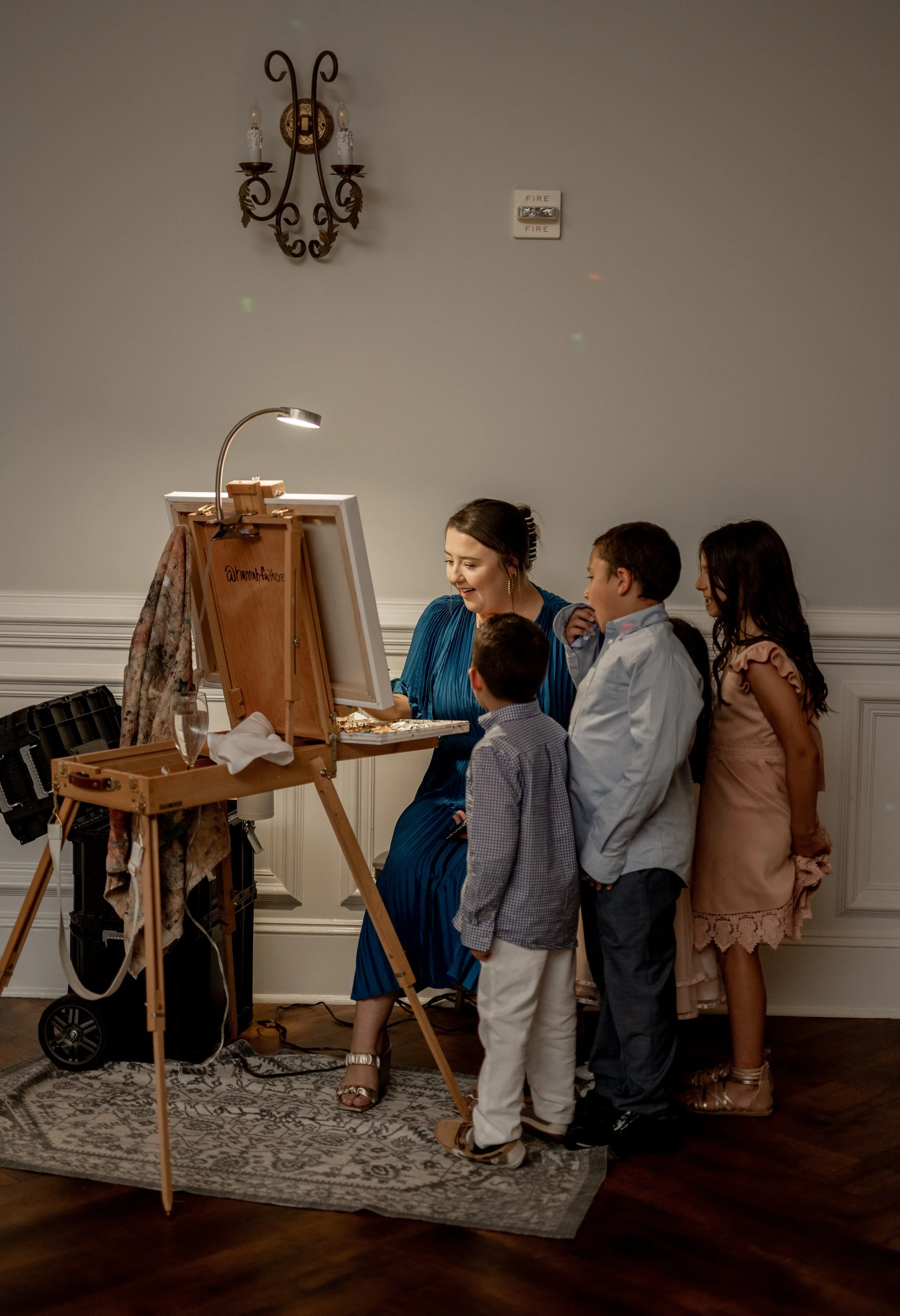 A woman is performing paintings or drawing on an easel surrounded by four children who are watching her work.