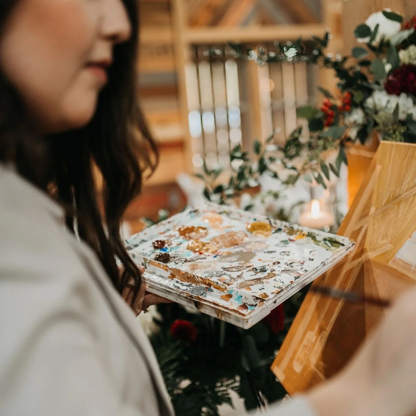 A woman holding a paint palette while painting, with an easel and flowers in the background.