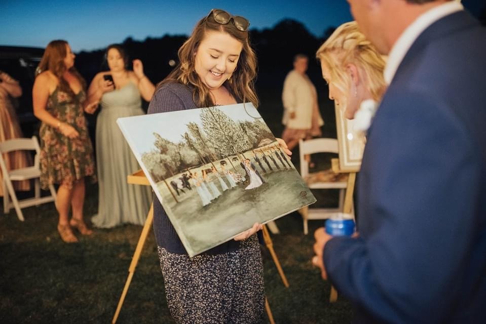 A woman in a gray sweater and floral skirt shows a painting to a man in a blue suit at an outdoor gathering during the evening. Other people are in the background, some taking photos, with chairs and artwork displayed around.