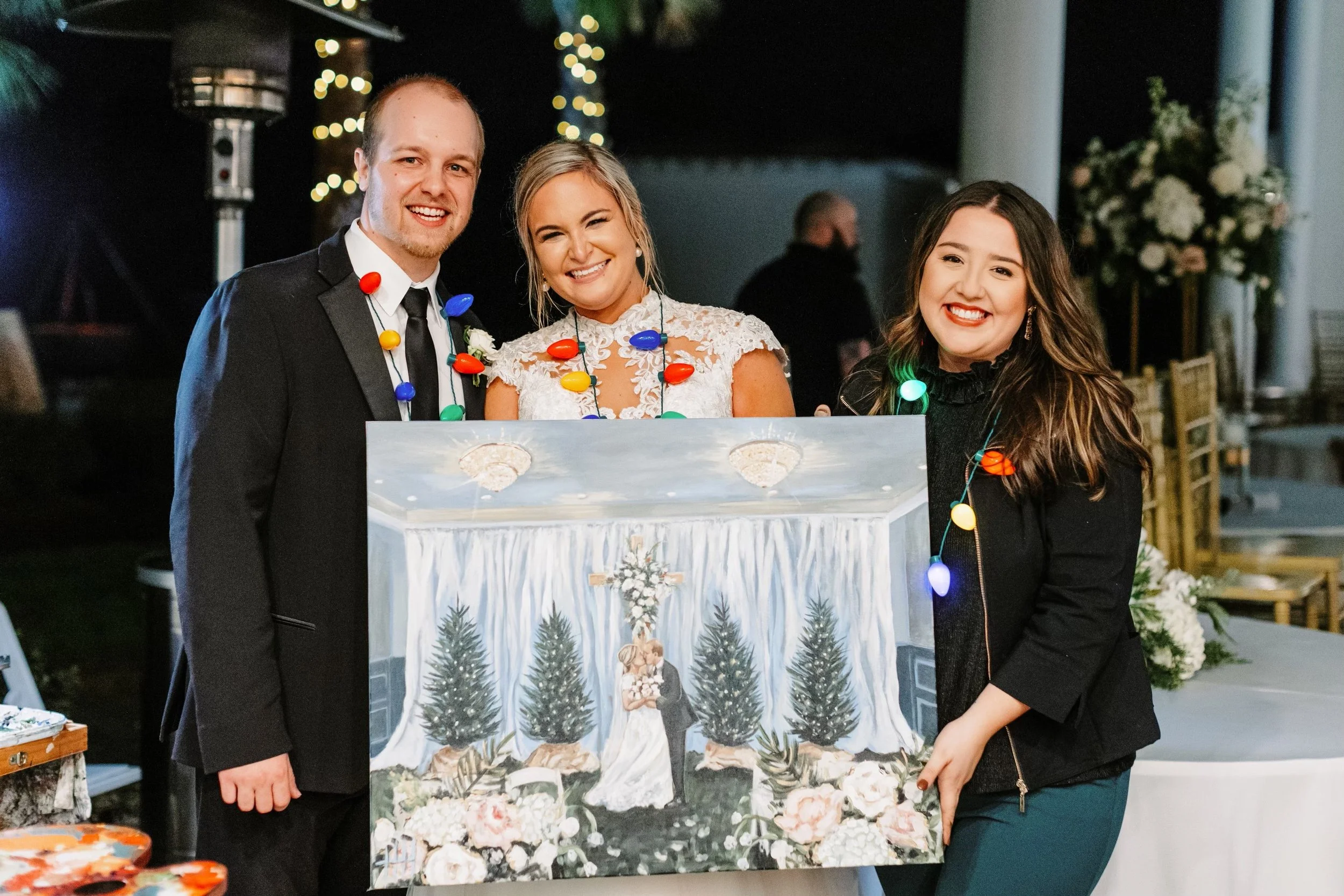 Three people smiling and holding a painting of a wedding scene, at a festive event decorated with string lights and flowers.