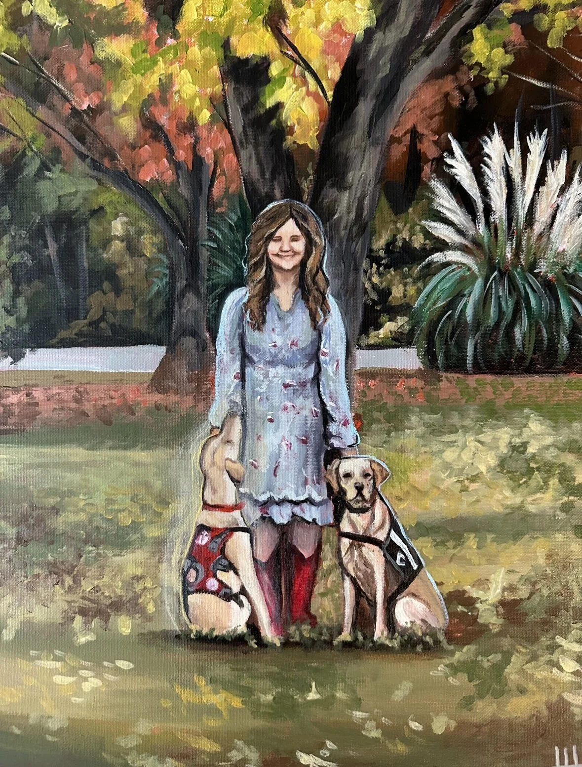 A woman with long hair smiling in a park during autumn, flanked by two dogs, one smaller dog wearing a polka-dot outfit and a larger dog wearing a harness, both sitting on the grass in front of a large tree with colorful fall leaves.