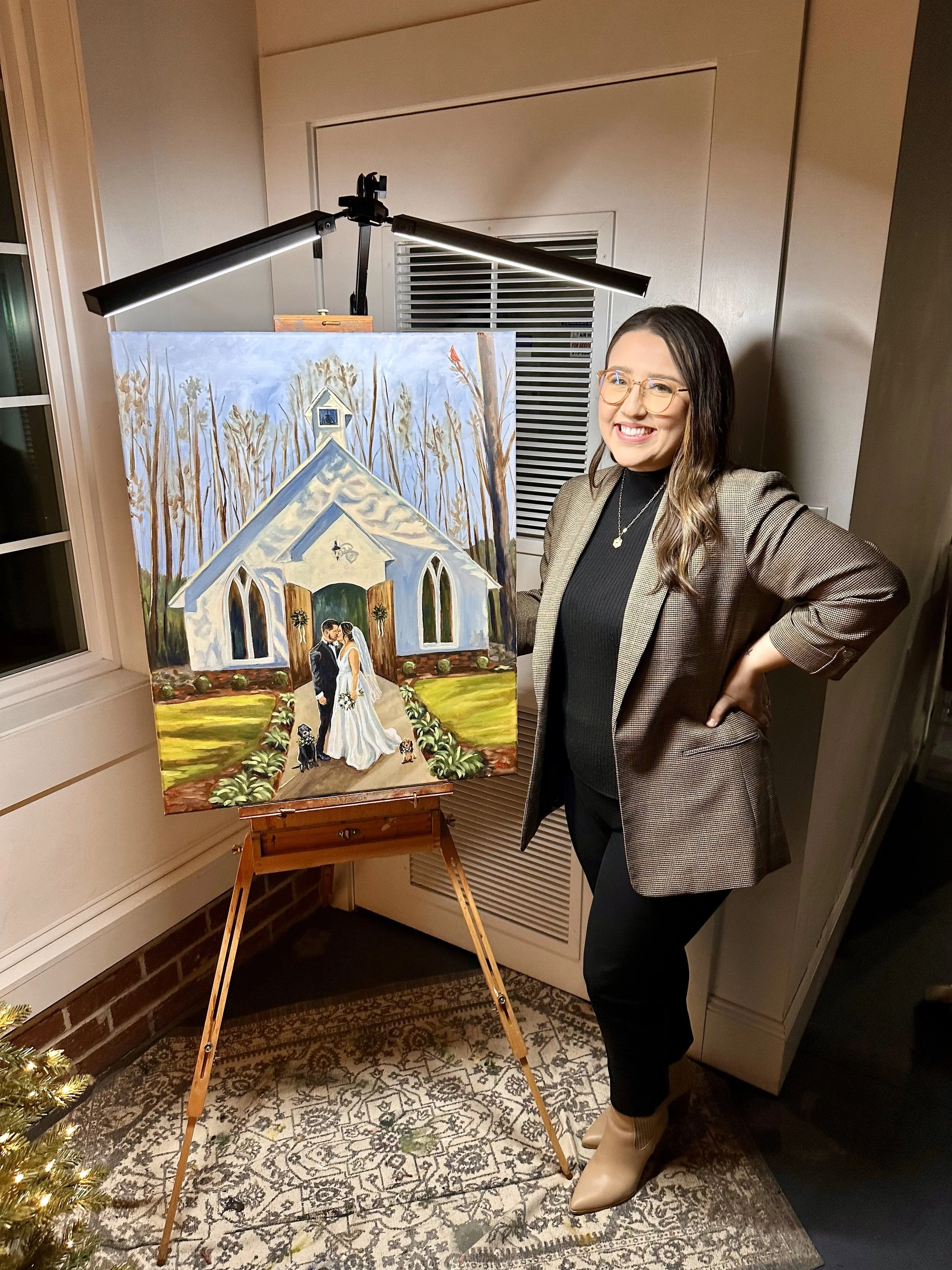 A woman wearing glasses and a blazer stands next to a colorful painting of a wedding scene in front of a church, smiling at the camera.