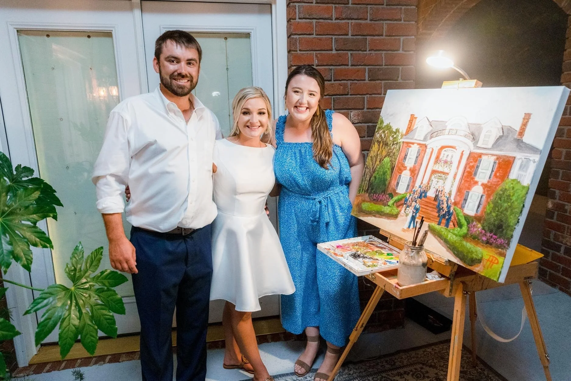 Three people smiling and posing for a photo indoors near a brick wall. A man in a white shirt and dark pants is on the left, a woman in a white dress in the middle, and a woman in a blue dress on the right. They are standing next to a large painting 