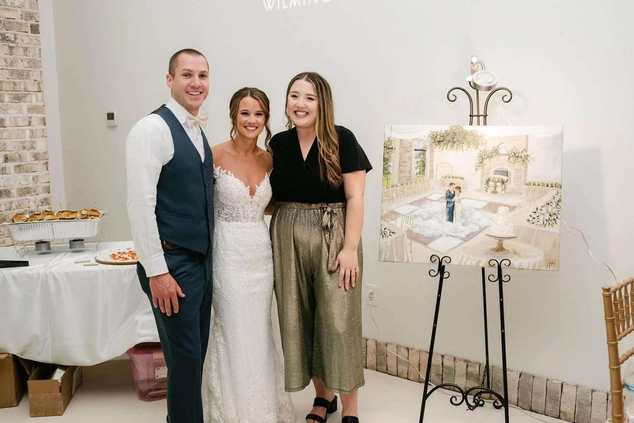 A bride in a white wedding dress stands between a groom in a suit and a woman in gold metallic pants, all smiling and standing indoors at a wedding celebration.