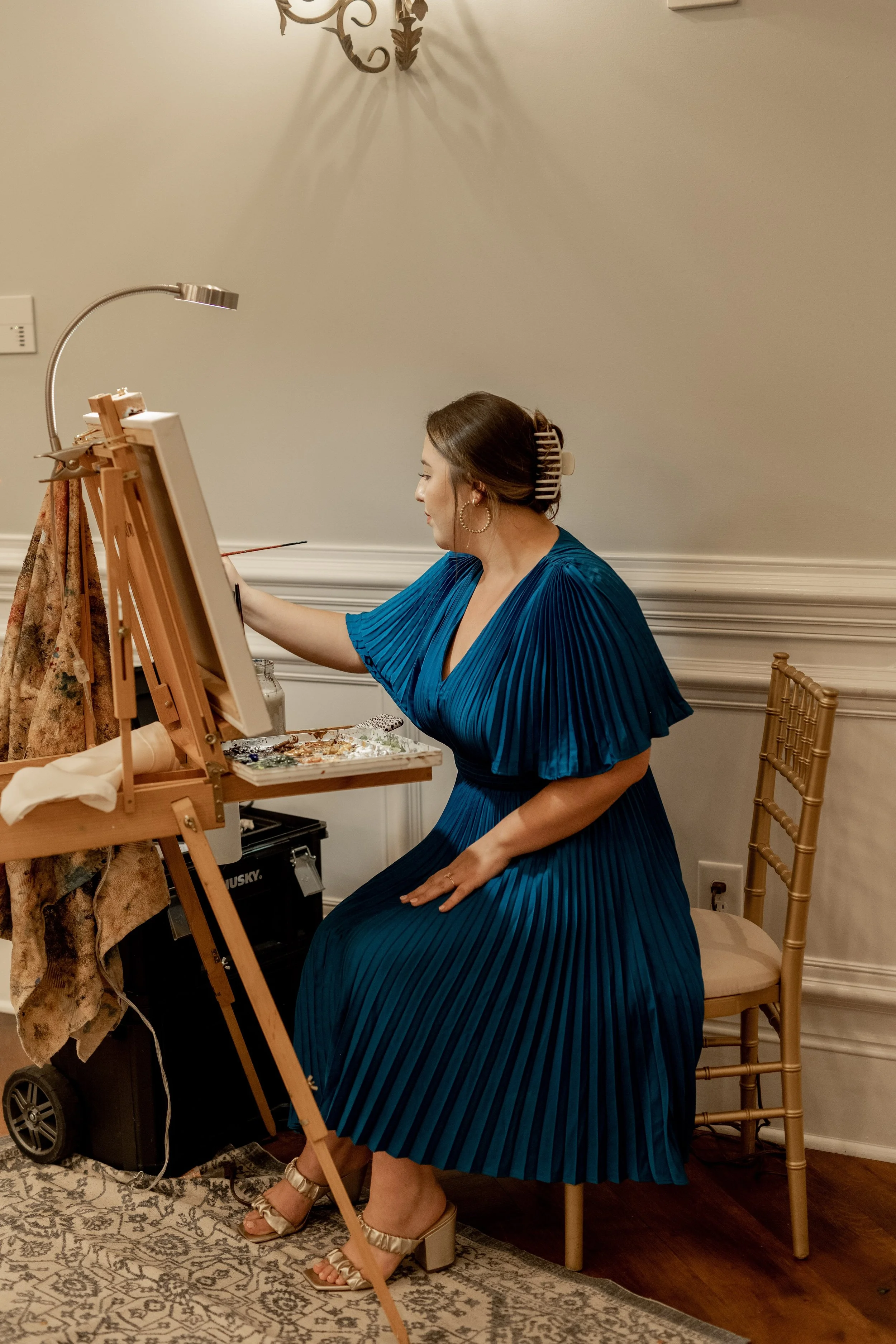 A woman in a blue pleated dress is seated on a chair, painting on an easel in a room with white walls and a decorative wall sconce.