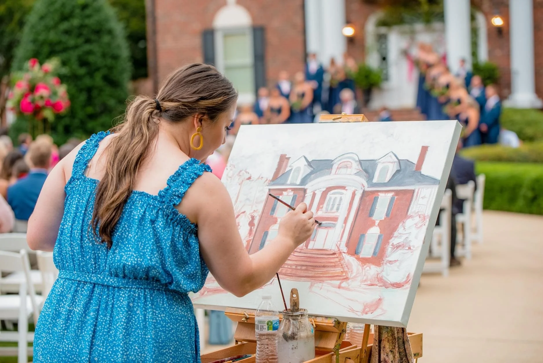 Woman in blue dress painting a portrait of a mansion on an outdoor canvas with an audience seated behind, at a formal event or wedding.