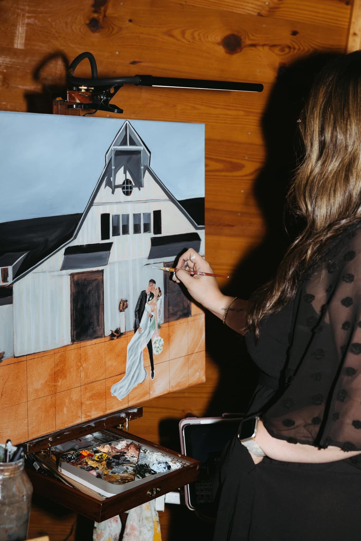 A woman painting a wedding scene on a canvas, with a barn in the background, using a palette of paints on a table nearby.