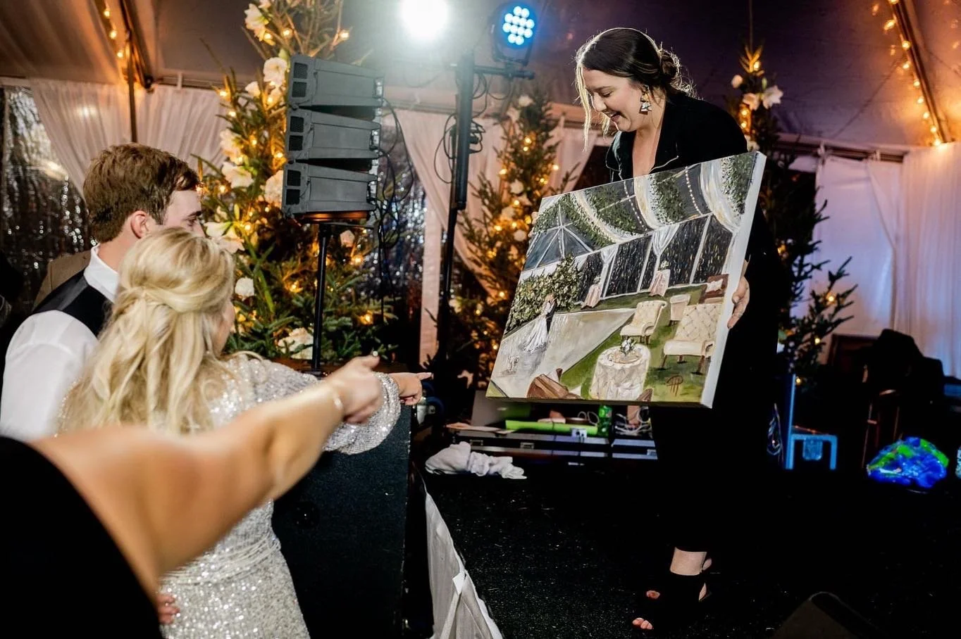 A woman presenting a painting of an outdoor garden scene to two guests at a festive event. The scene features a patio with chairs and greenery. The background has Christmas trees, string lights, and a tent.