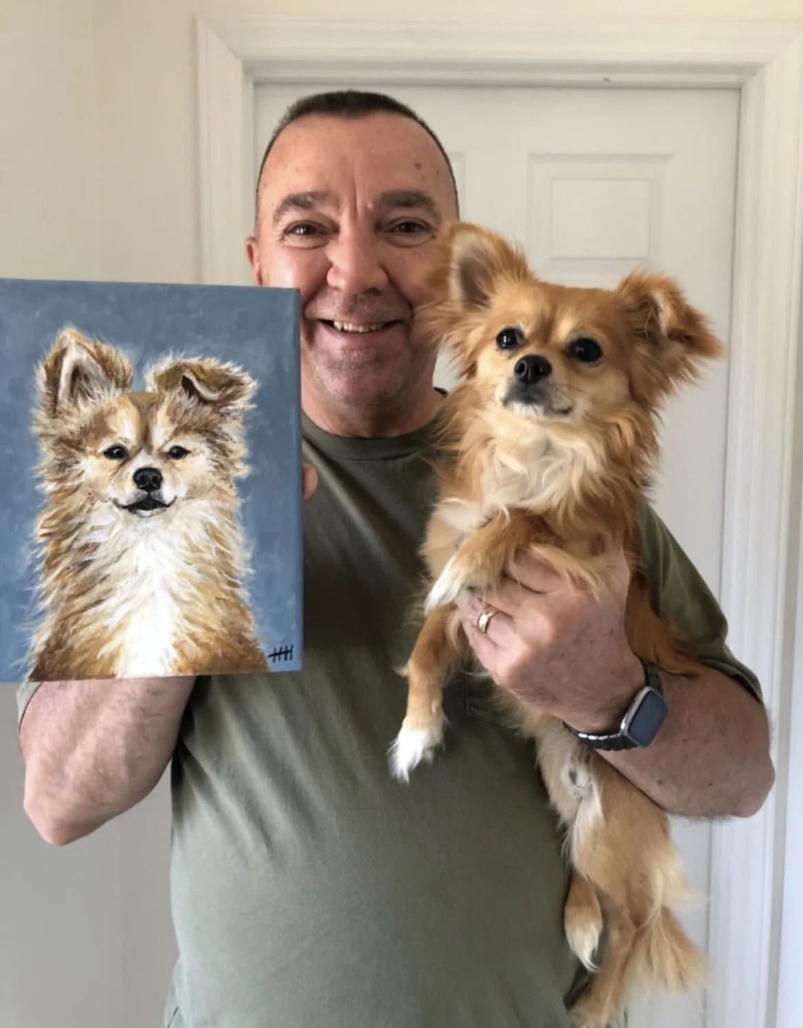 A man smiling and holding a small tan dog with fluffy ears and a white chest, while displaying a painting of a similar dog.