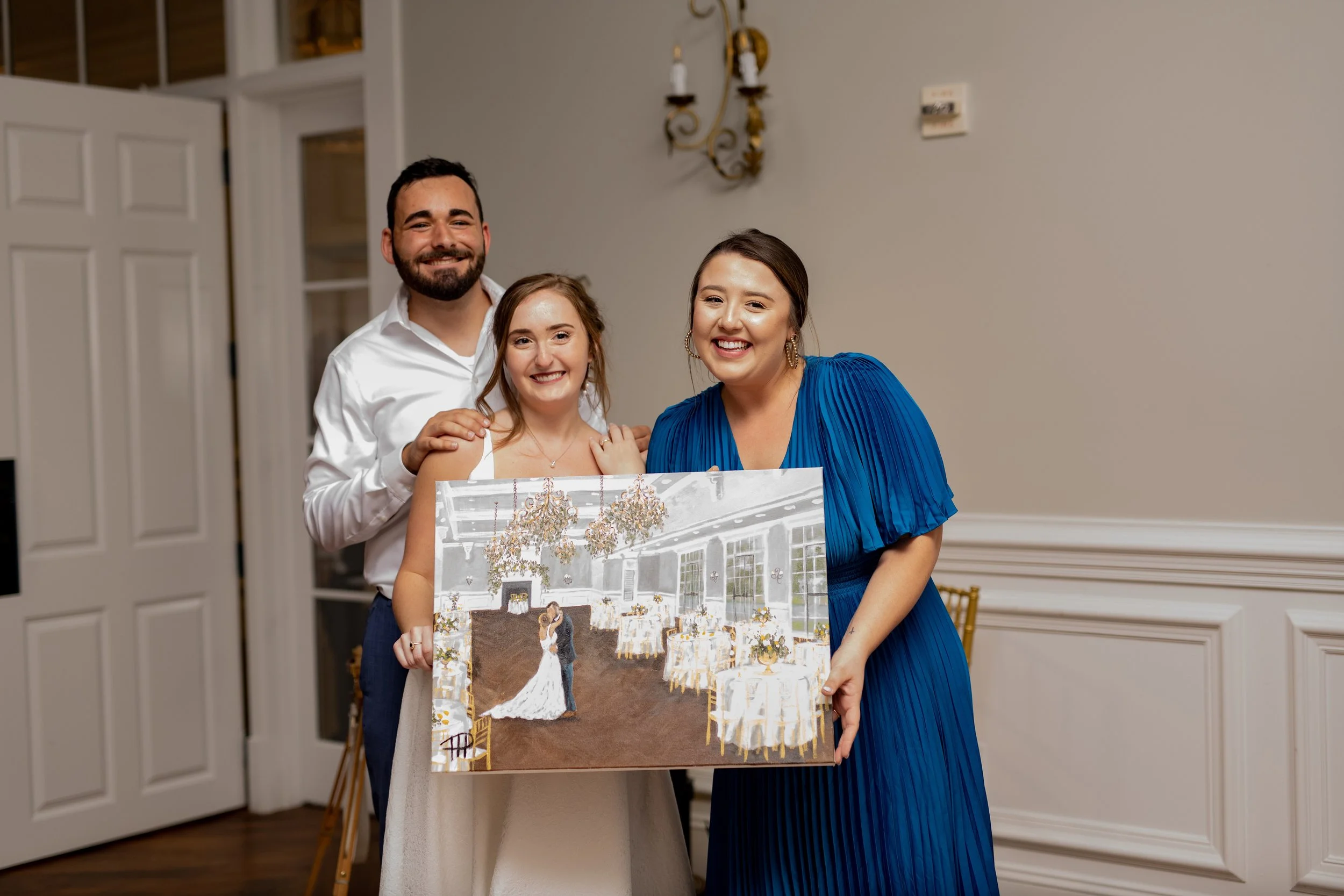 Three people smiling, including a woman in a white dress holding a wedding painting, in a decorated event space.