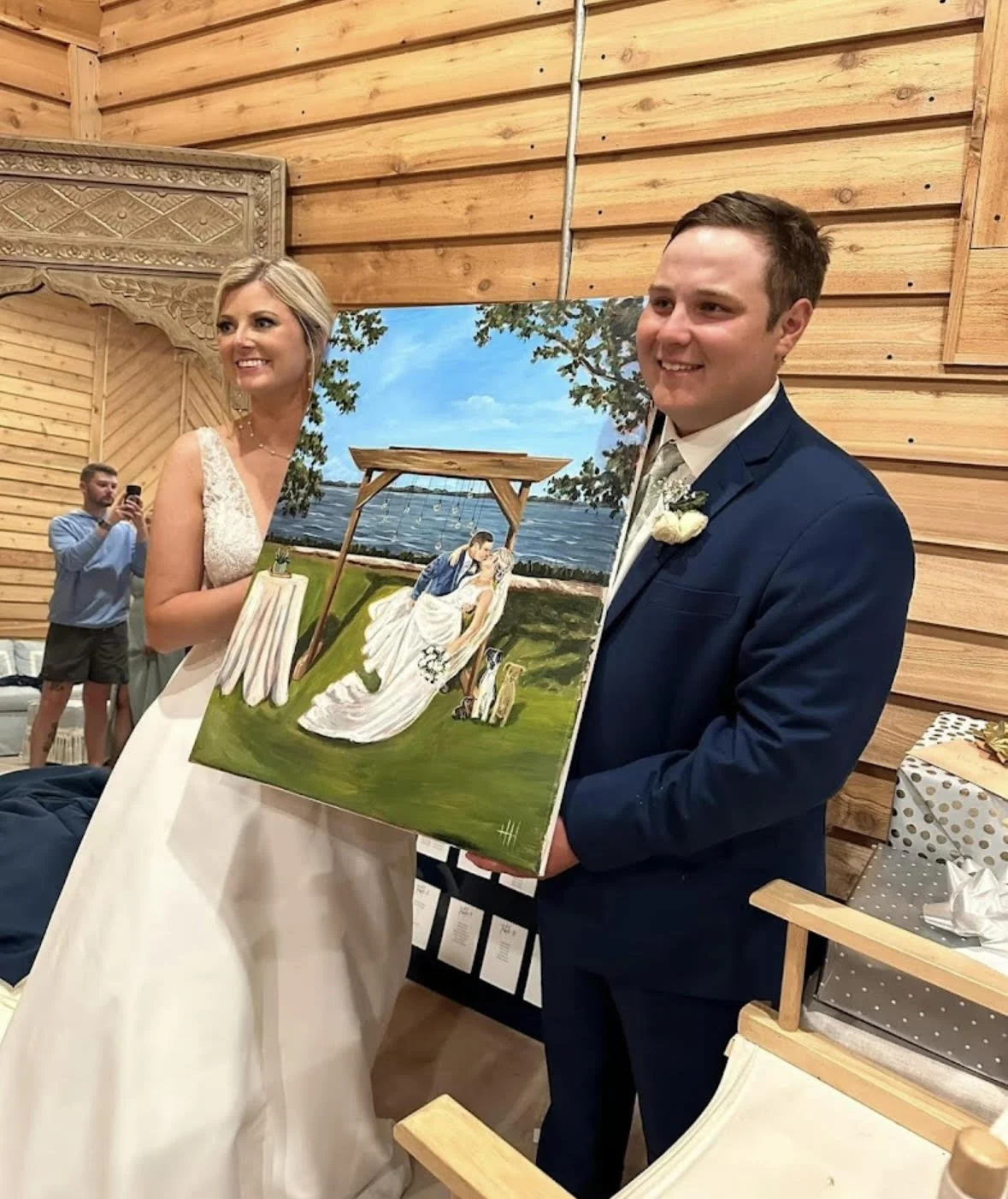 A couple dressed in wedding attire holding a painting at a wooden event space. The painting depicts a wedding scene by the water with a bride and groom under a wooden arbor.