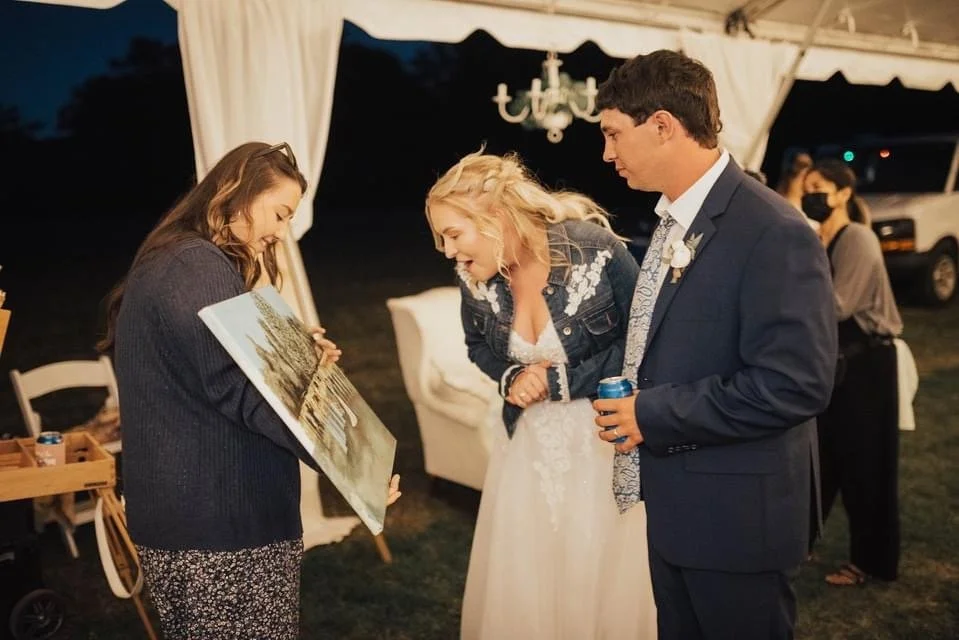Three people, two women and a man, stand together at a celebration under a tent, looking at and smiling at a painting held by a woman. The woman in the middle is wearing a white dress and jacket, while the man is in a suit holding a beverage. The wom