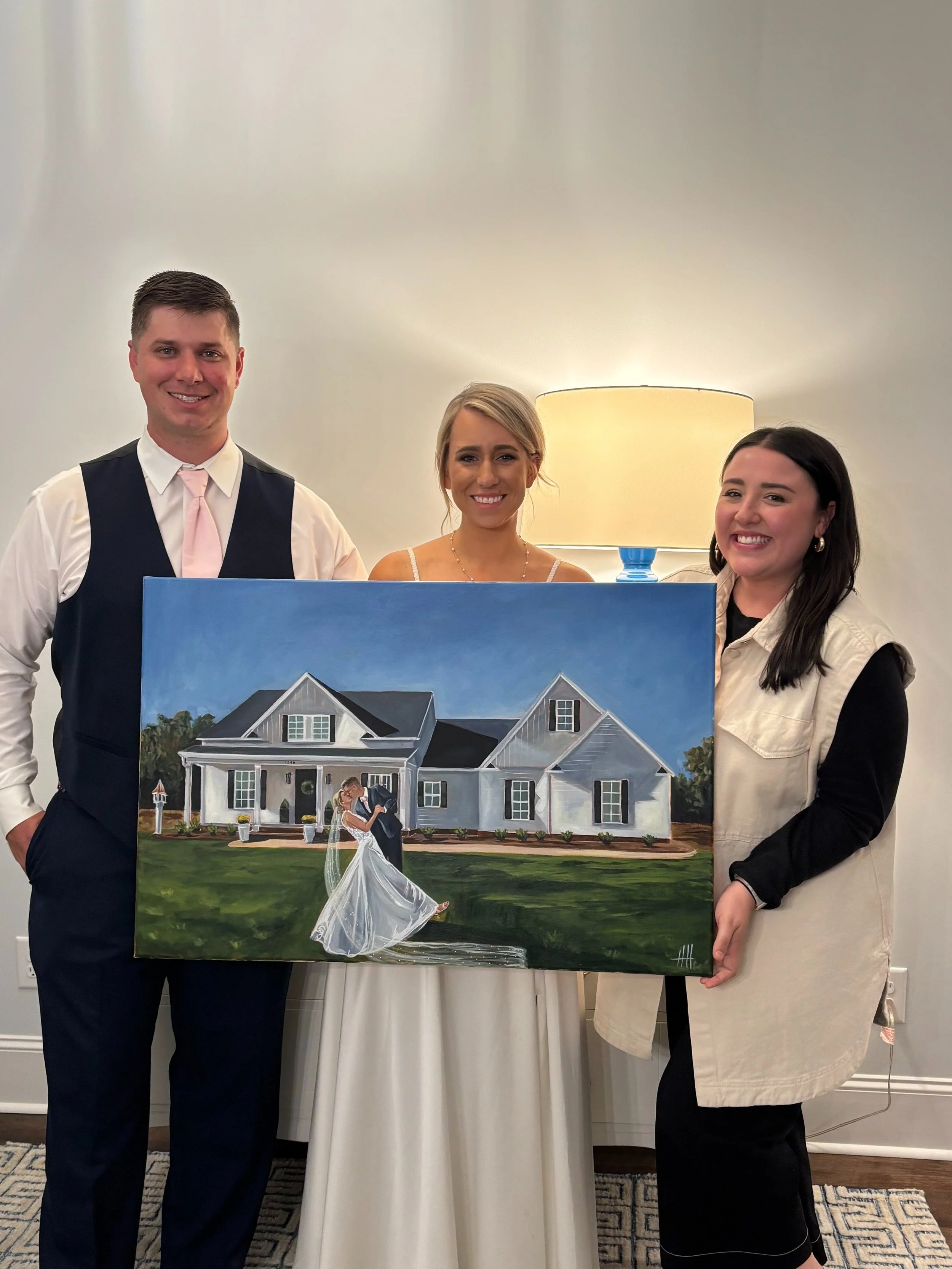 Three people standing behind a painting of a house and a dancing bride and groom, indoors with a lamp in the background.