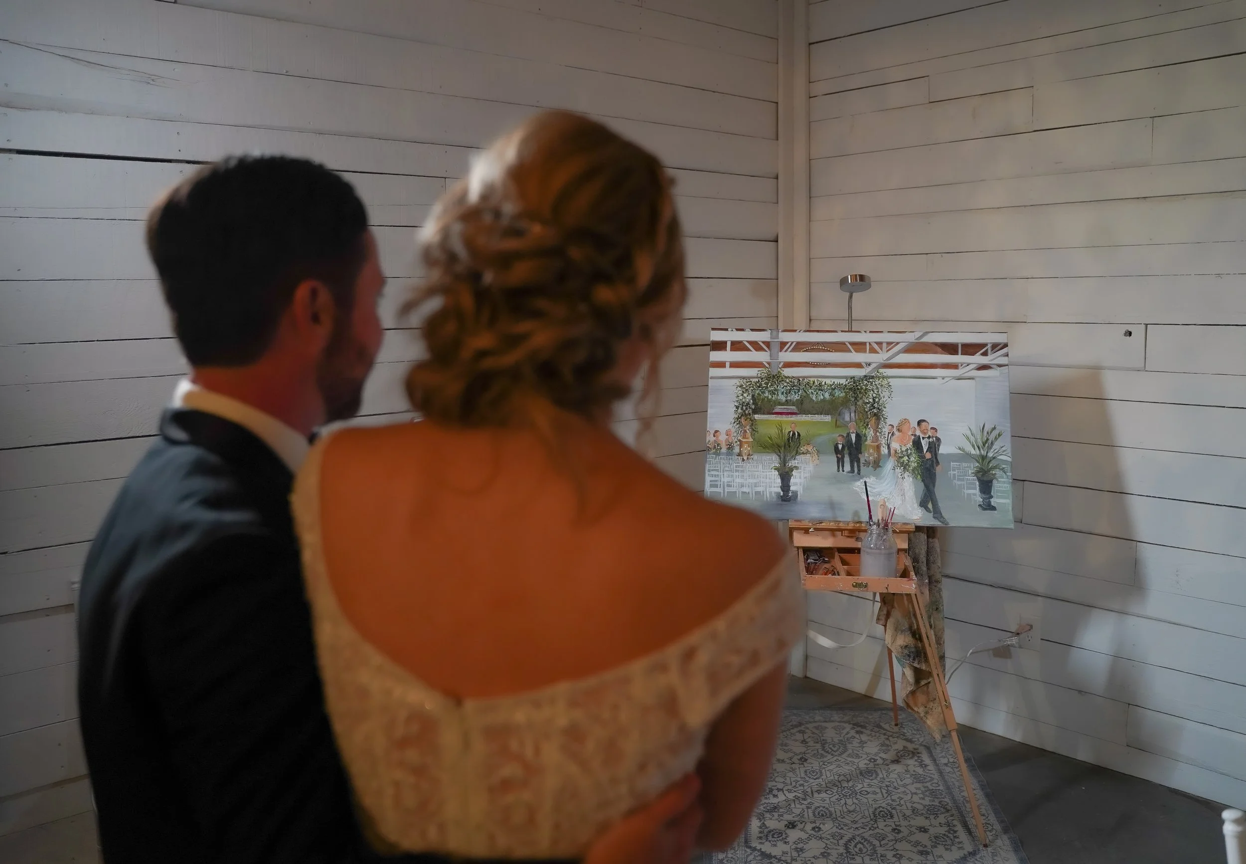 A bride and groom watching a large painting of a wedding scene inside a white-washed wooden room.