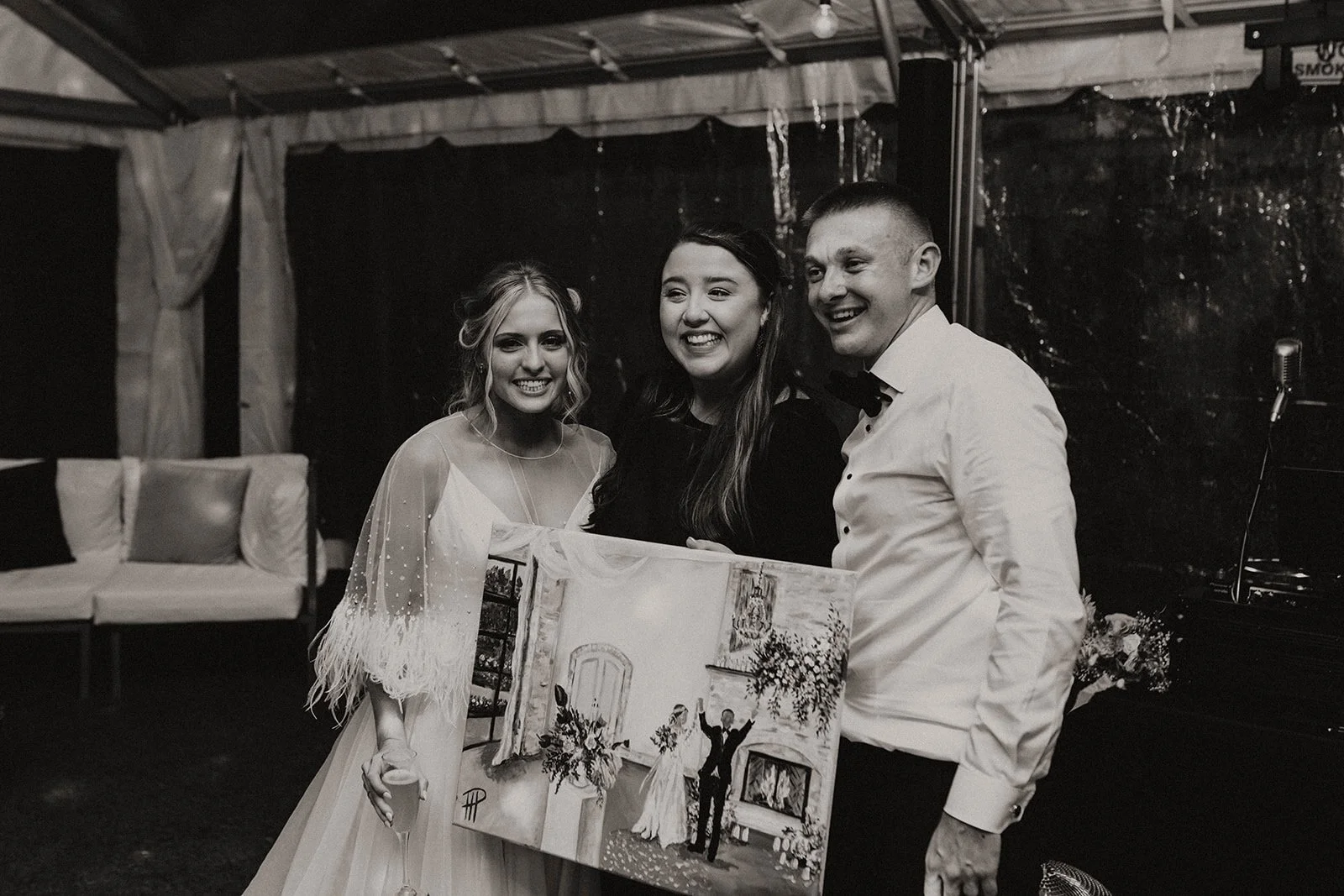 Three people smiling at a celebration, holding a painting in a decorated indoor venue.