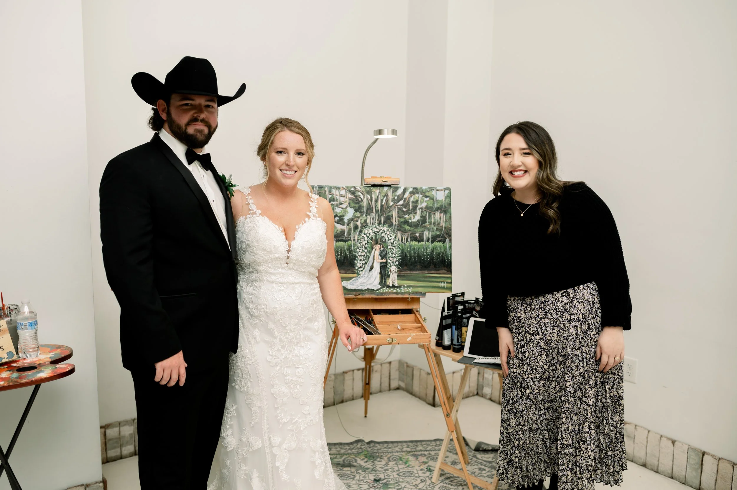 A man in a tuxedo and cowboy hat, a woman in a wedding dress, and a woman in a black sweater and patterned skirt standing together indoors with wedding artwork in the background.